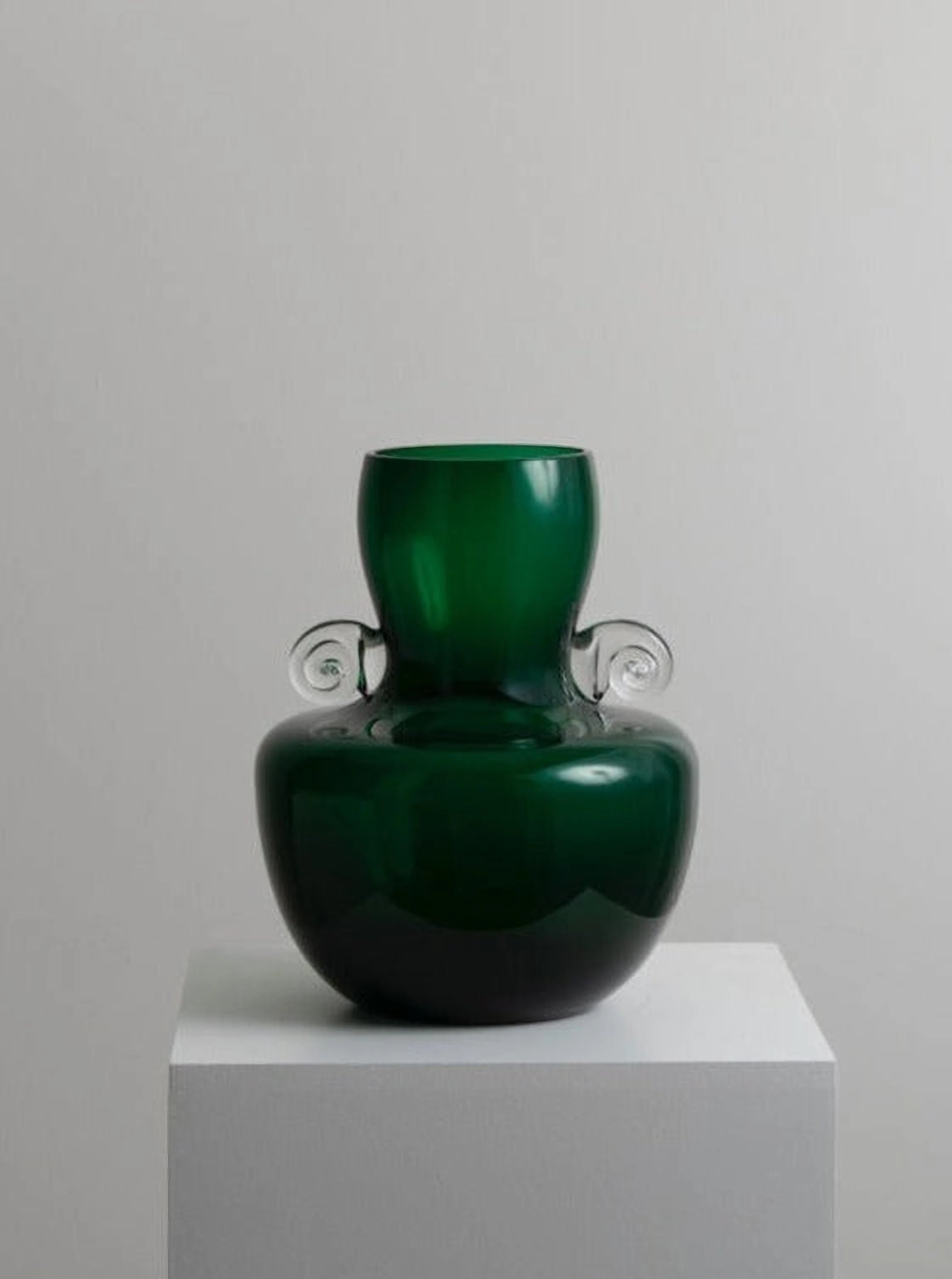A Vintage Bohemian Vase by Les Objets Amsterdam in mint condition, with a green hue and rounded base, adorned with two clear, spiral-shaped handles, sits on a white pedestal against a plain gray background.