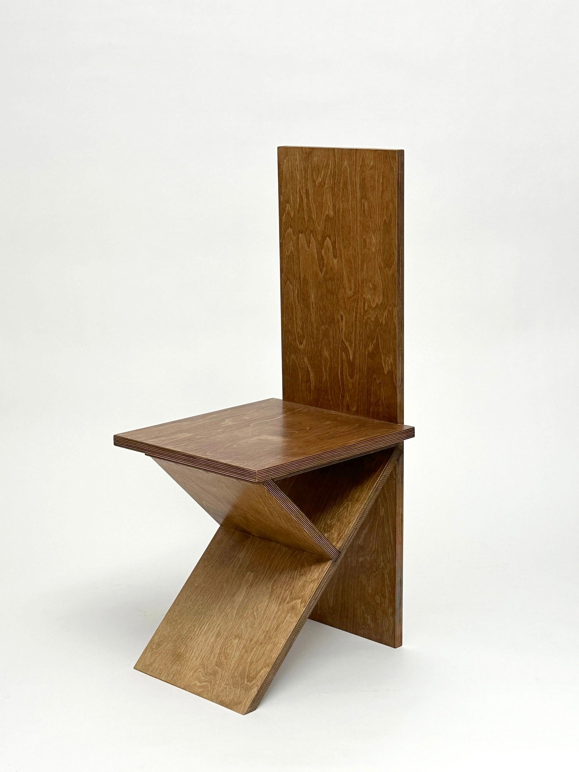 Introducing the Flat Pack Chair by GOONS: a distinctively crafted wooden dining chair with geometric lines. It showcases a tall, rectangular backrest and a seat that seamlessly intersects at an angle with the base. This artisanal masterpiece is handcrafted to deliver an abstract, modern aesthetic, perfectly contrasted against a simple, light-hued backdrop.