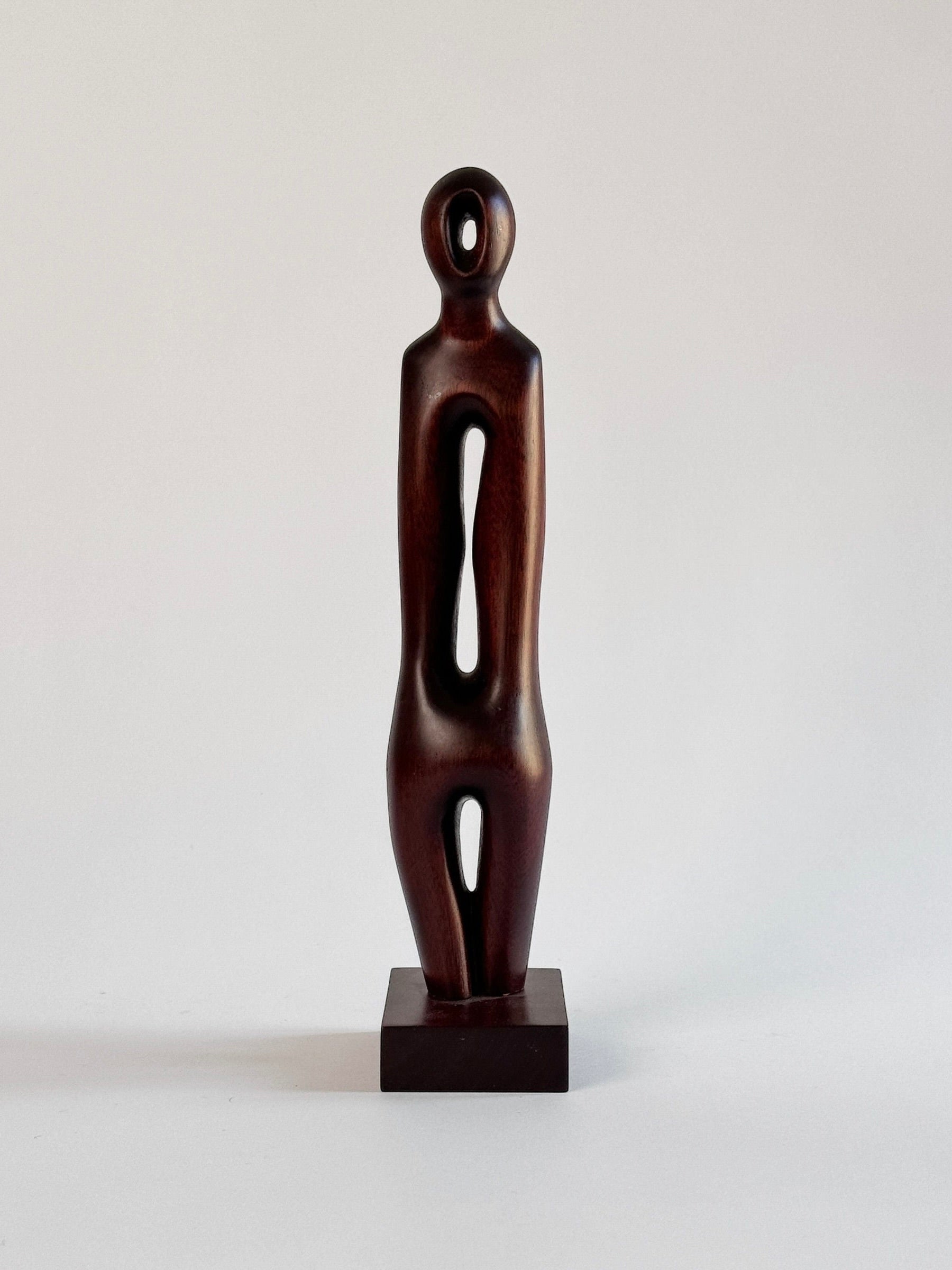 The Exotic Wood Sculpture by Pierre Dedieu from Figuresdesign is a minimalist piece featuring a human figure with a cylindrical head, elongated body, and stylized cutouts. This unique sculpture stands on a square base, showcasing the smooth and polished dark wood grain against a plain, light background.