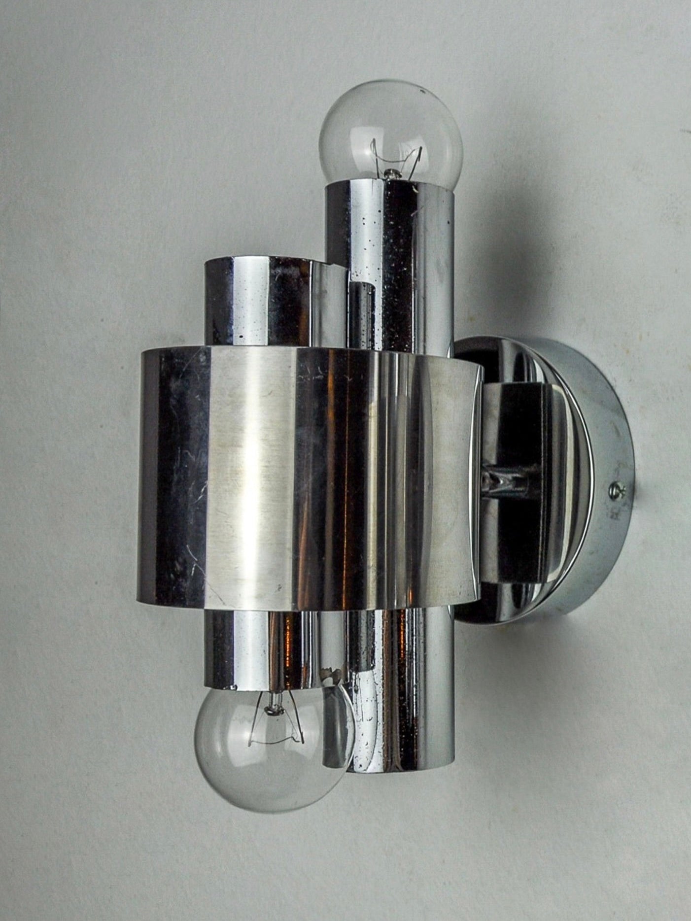A contemporary Geometric Chromed Sconce by Malamar Studio, dating back to Italy in 1970, showcases a sophisticated chrome-plated design with dual cylindrical components and two exposed round light bulbs. It is elegantly displayed on a plain white wall.