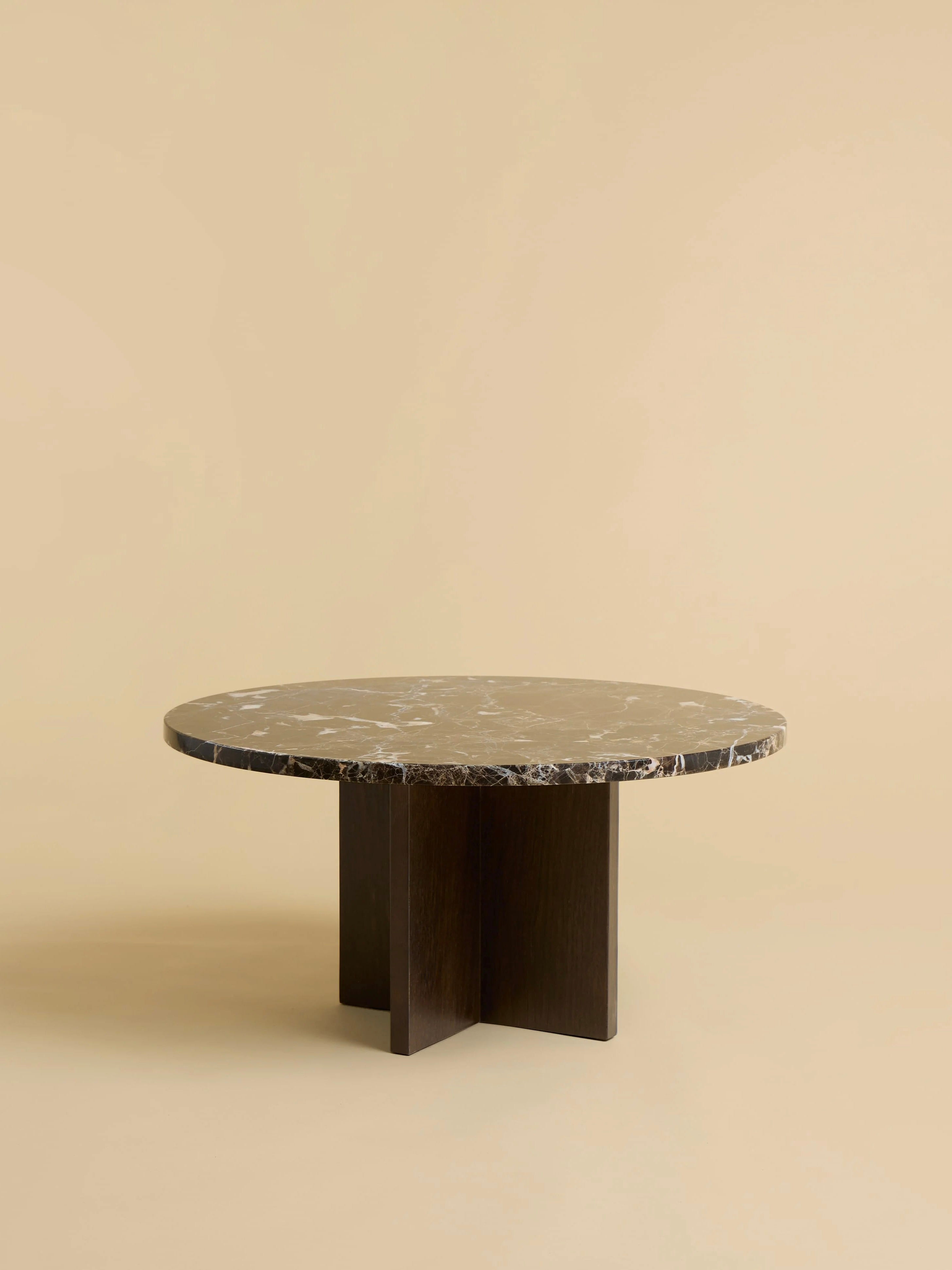 The "Tinian" Coffee Table by Lebanto features a round marble top with a natural pattern, resting on an artisan-crafted cross-shaped oak base, set against a plain beige background.