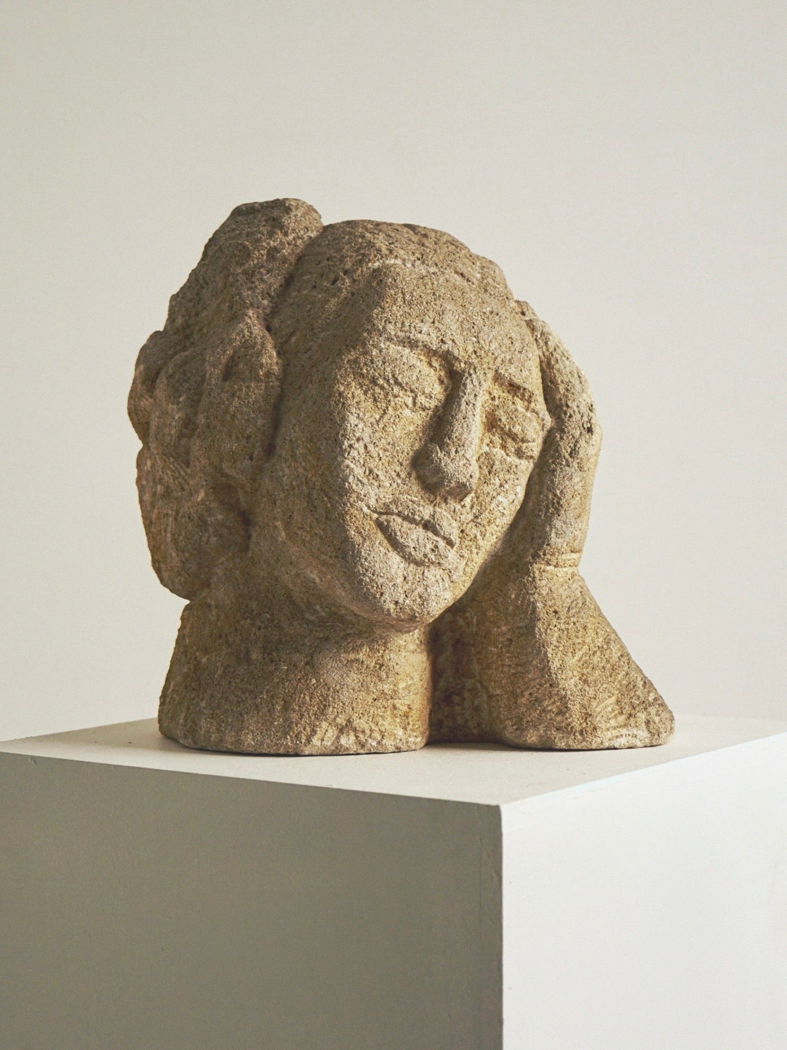 An Antique Carved Stone Bust of a Woman by Figuresdesign, featuring a serene textured face with closed eyes resting in her hand, reminiscent of an antique bust, is displayed on a white pedestal against a plain light wall.