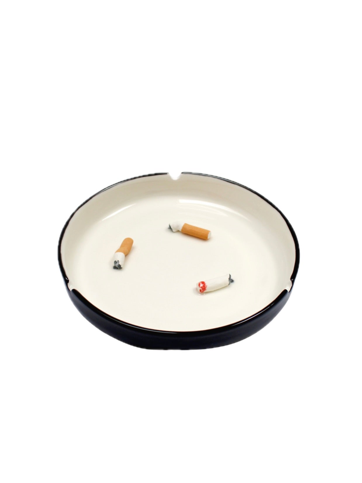 The Villa Arev Cigarettes Ashtray, crafted from high-quality earthenware with a black rim, contains three cigarette butts. This handmade white ashtray features edge grooves for holding cigarettes. The plain white background evokes the charm of a Parisian café scene.