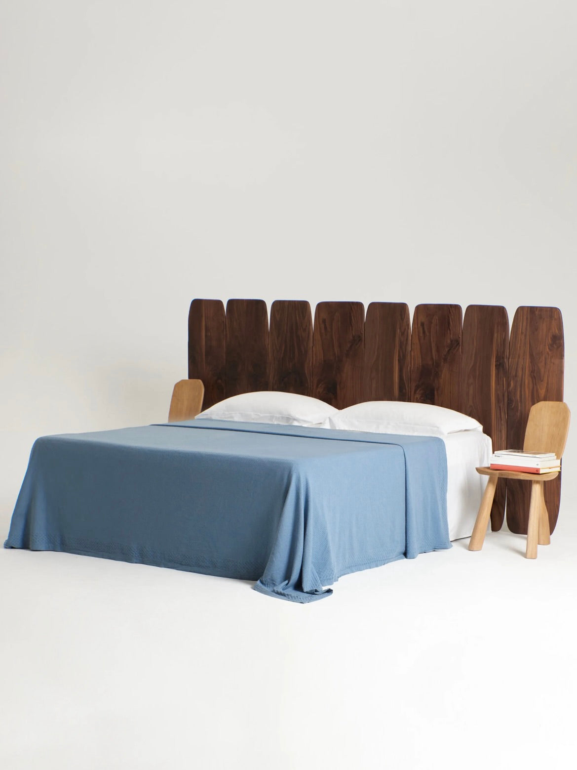 The stylish bed showcases the Project 213A Madeira Walnut Headboard Medium, handcrafted from high-quality walnut wood in Portugal and featuring individual vertical slats. It is decorated with a blue blanket and white pillows. To the right, a small wooden nightstand holds a pink book and tablet against a plain white background.