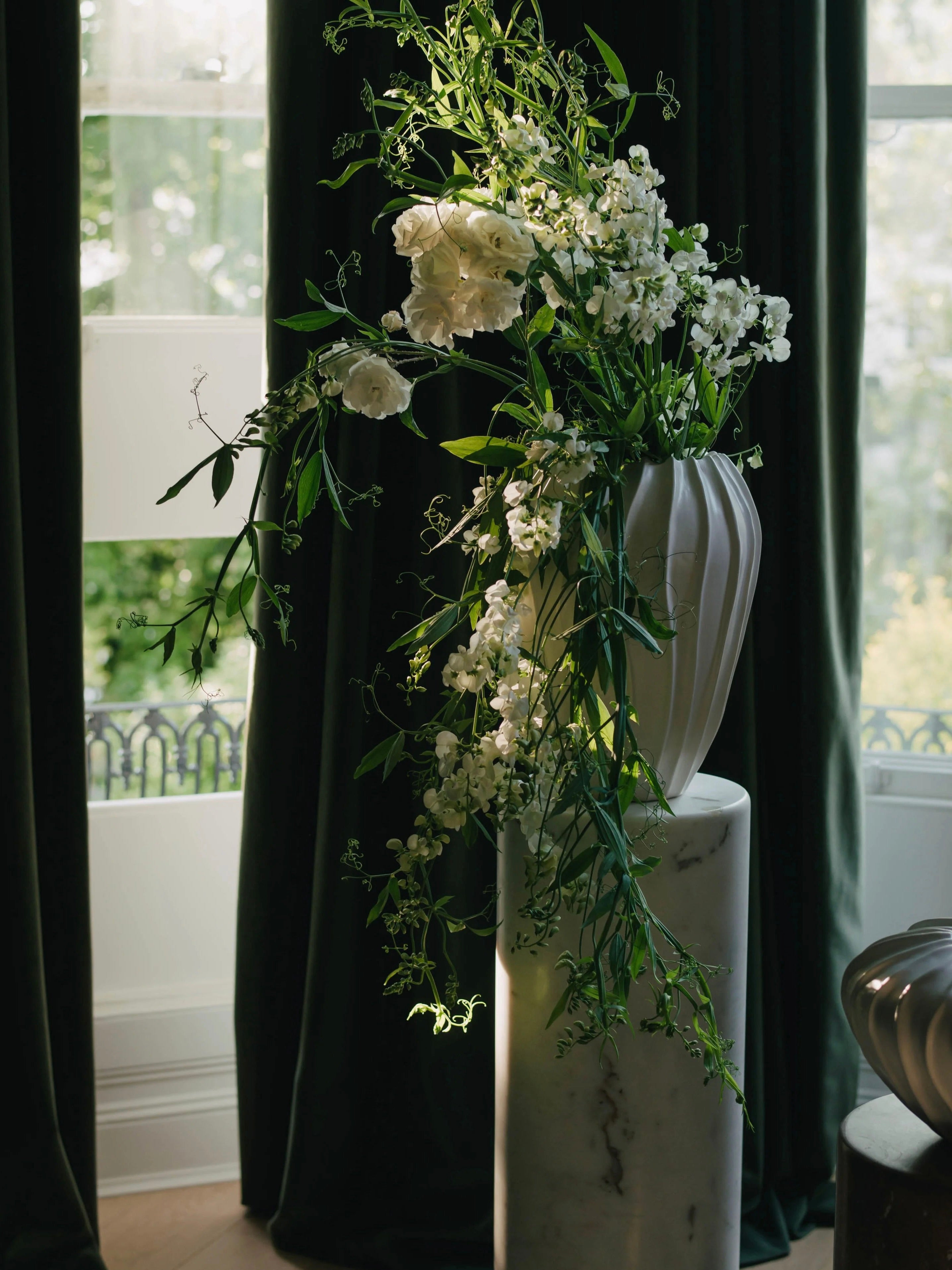 A tall, elegant Large Cloak Vase by Ransom & Dunn from the ceramic collection, handmade in Portugal, is filled with cascading white flowers and lush green foliage atop a marble pedestal. The scene is softly lit, with dark curtains and sunlight filtering through a window.