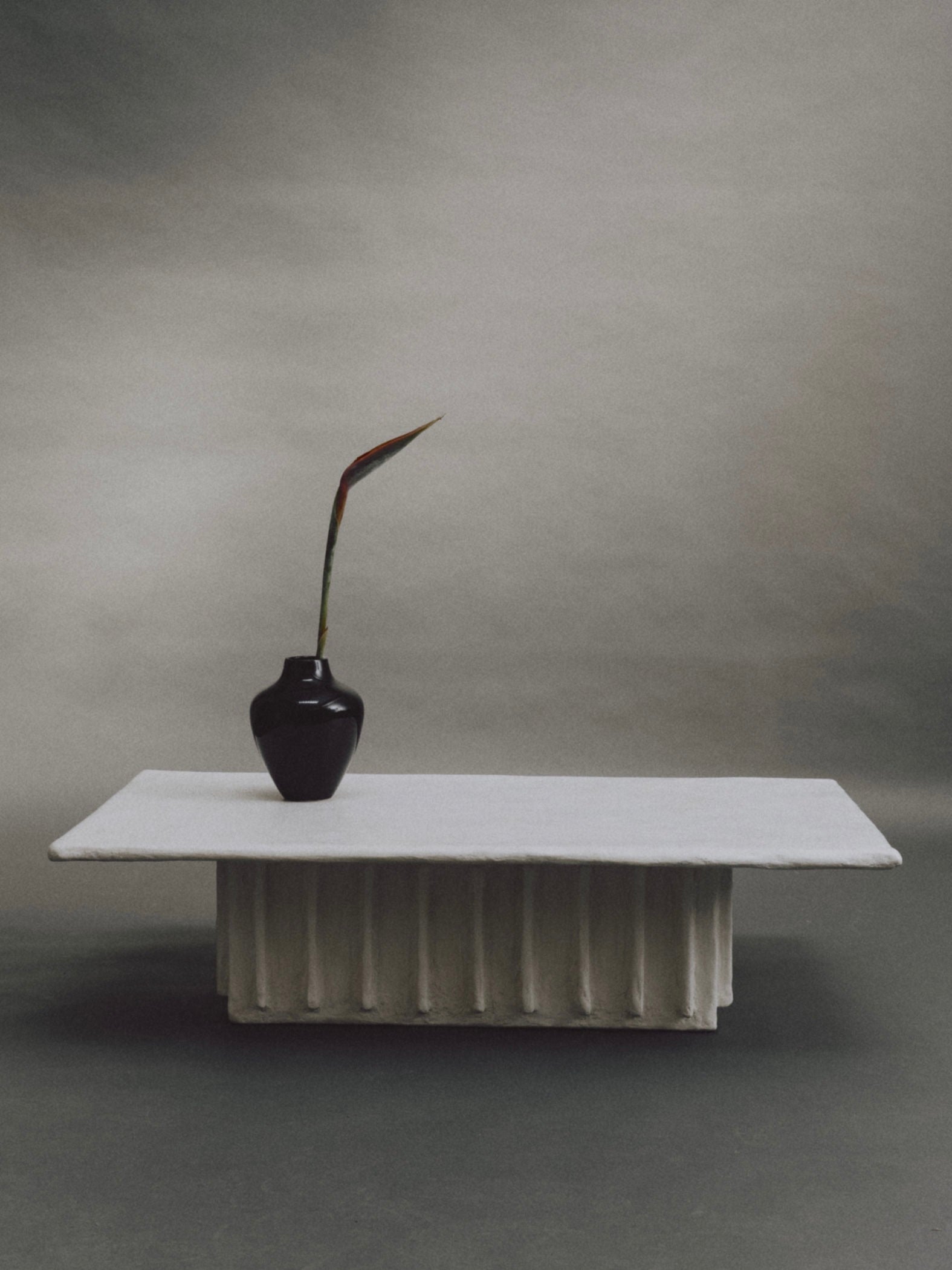 Experience minimalism with the Columnar Coffee Table by Ombia Studio: a black vase with a single long-stemmed leaf on a white, textured rectangular pedestal against a muted gradient background. Available in custom sizes and made to order for your space.