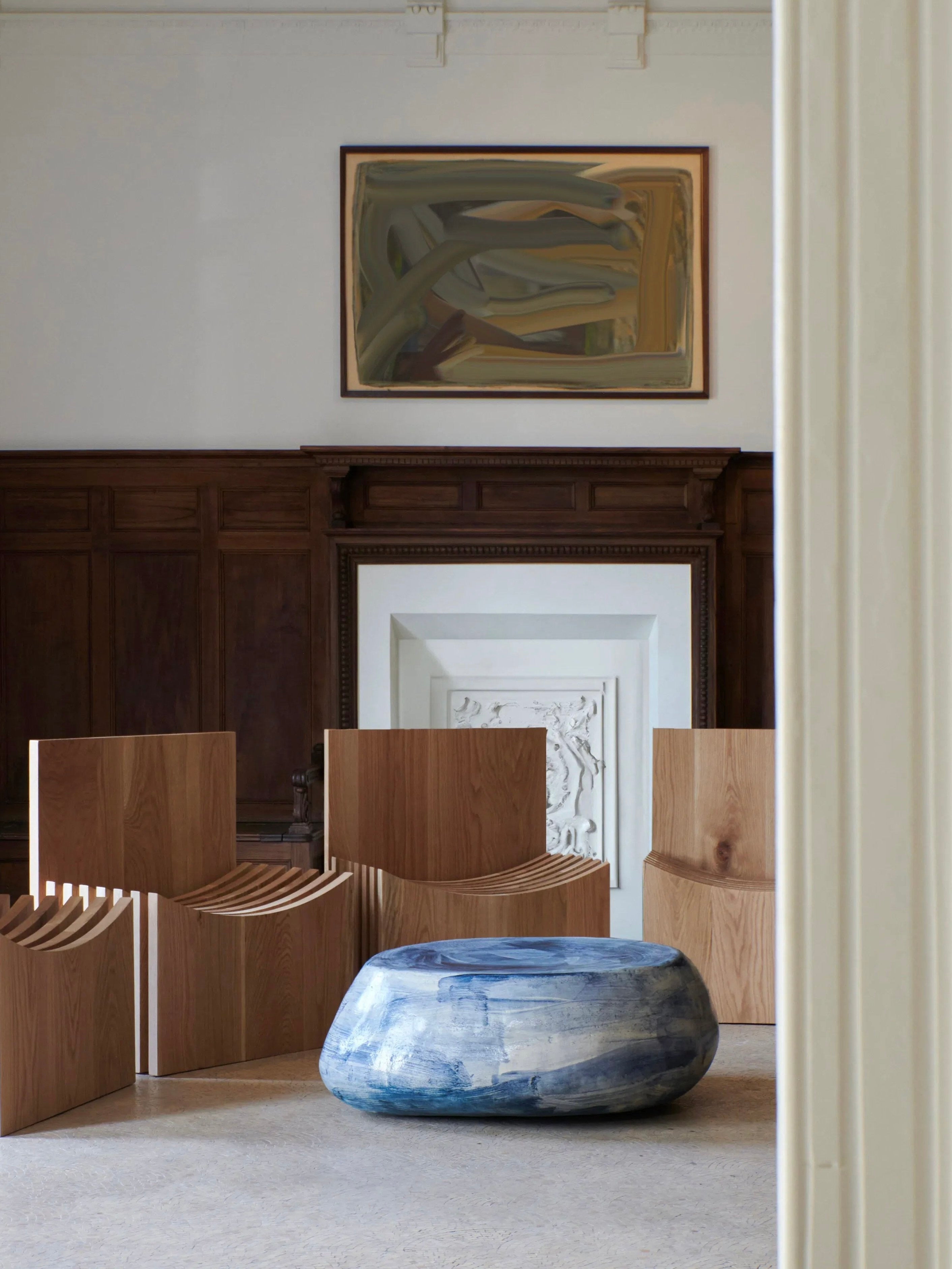 A modern, minimalist room features wooden slatted chairs and a round, Brushed Blue Ceramic Coffee Table from Project 213A. The space is adorned with dark wood paneling, a white ornate fireplace, and a contemporary abstract painting above the mantel. The bespoke dimensions of each piece add to the room's unique character.