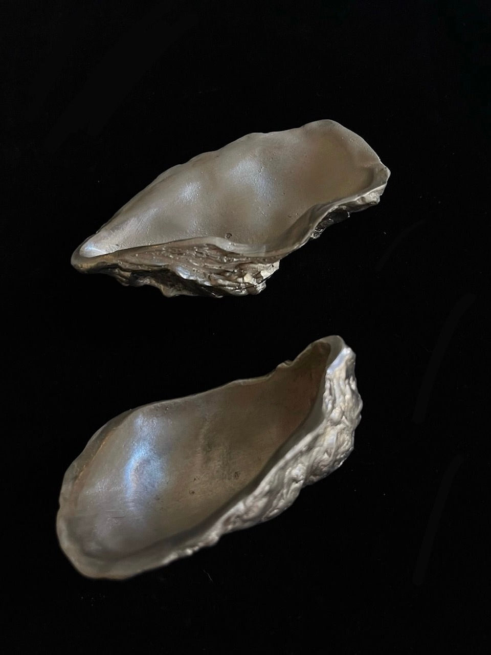 The Handcrafted Silver Oyster Set by Médecine, featuring two shells with smooth, curved interiors and rough, textured exteriors, lies against a black background like silver-plated treasures.