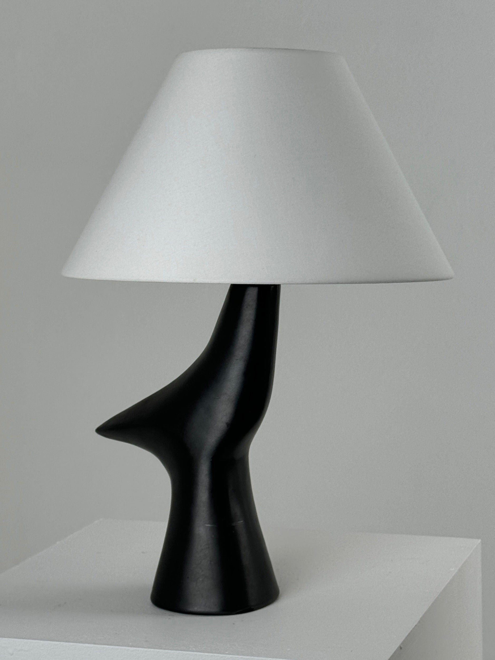 The Zoomorphic Ceramic Lamp by Figuresdesign features a sleek, sculptural black glazed ceramic base inspired by France 1960, complemented by a white conical lampshade. It is elegantly displayed on a minimalist white surface against a light gray background.