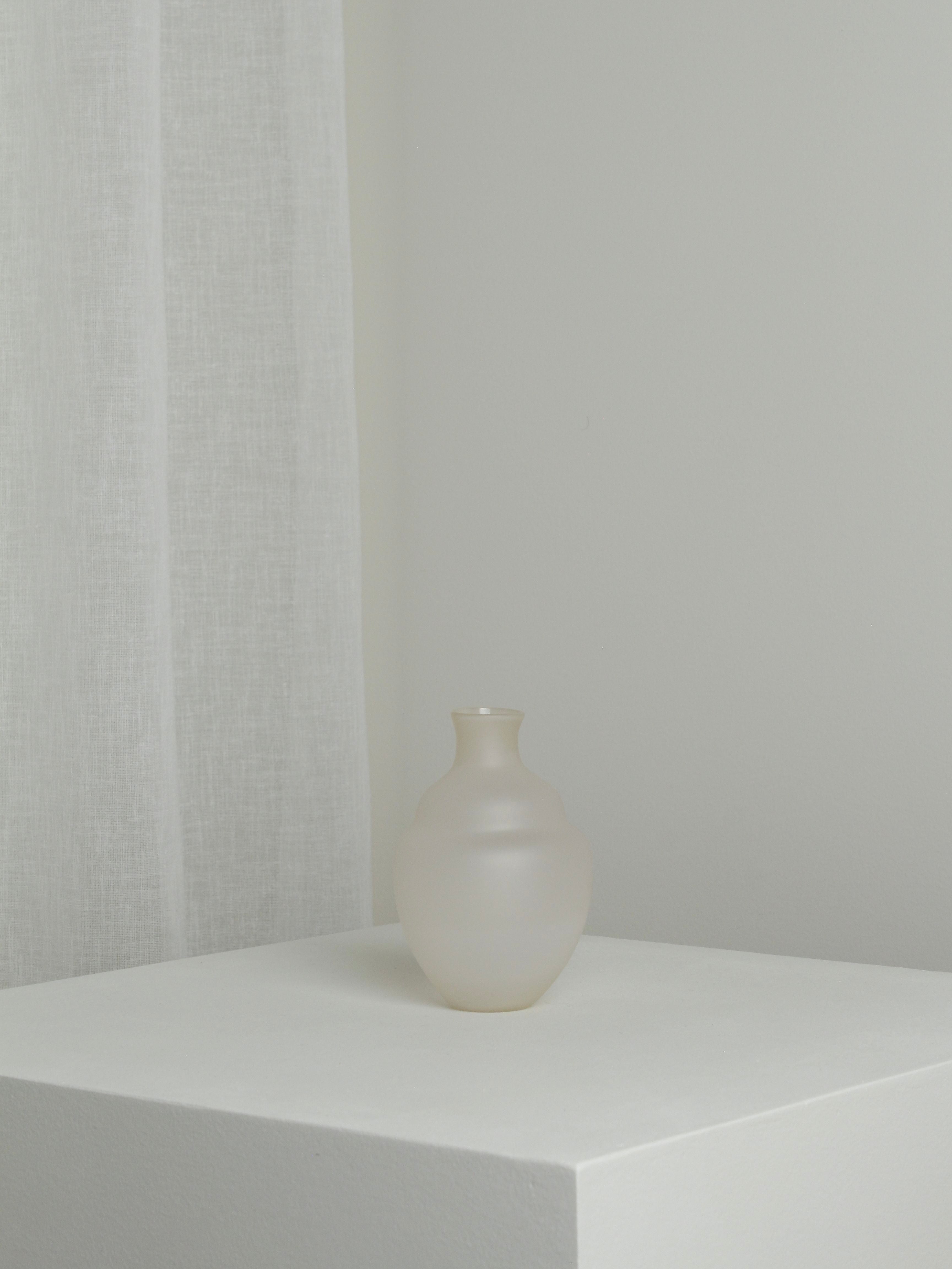 A Verre d'Onge Small Vase in Moon sits on a white pedestal against a backdrop of a white curtain, reminiscent of the calm night skies over Montreal. The scene is minimalist, with soft lighting that highlights the vase's smooth, rounded shape and the subtle texture of the curtain.