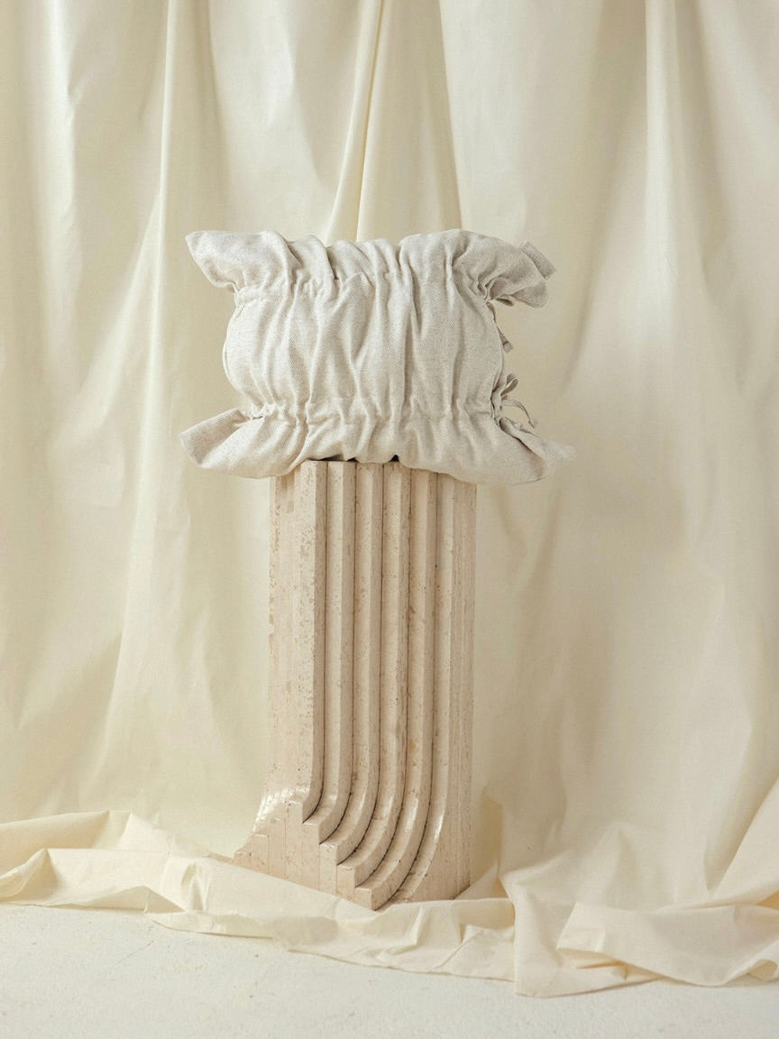 The Daphne Sand Cushion by SANTA LIVING, made from recycled polyester fibers and featuring ruffled edges, rests on a simple ribbed pedestal against draped cream fabric.