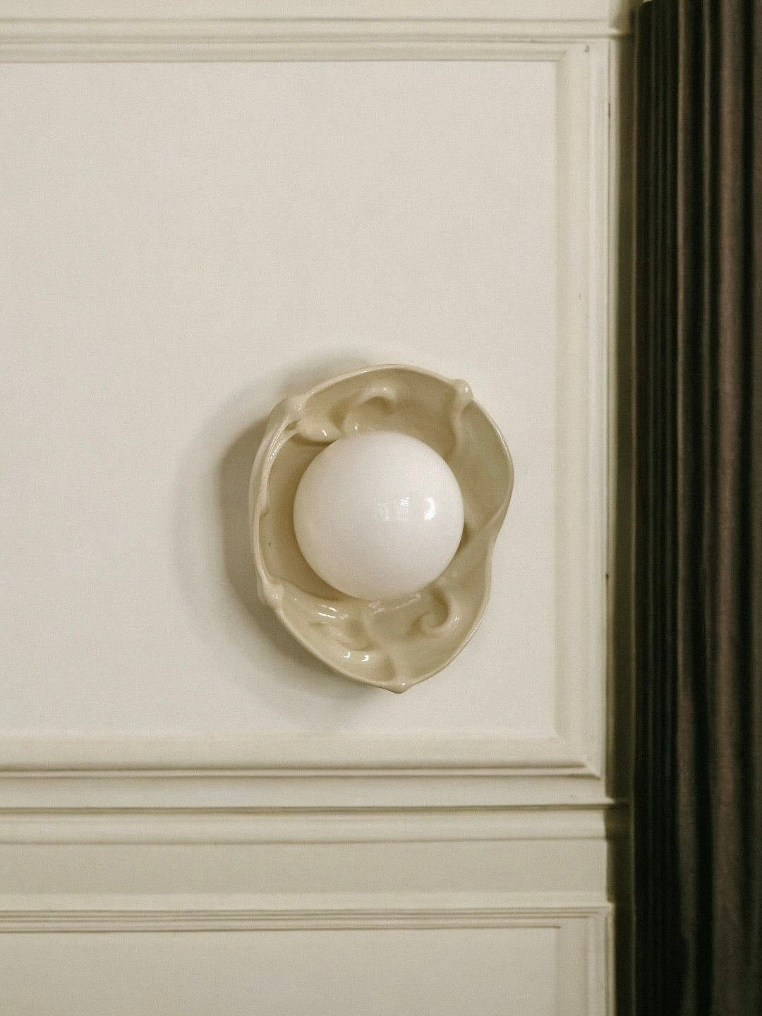 The 'Velour' Orb Wall Sconce by Gazar Studio, featuring a round white light and an irregular stoneware base, is mounted on a cream wall with decorative molding. A dark curtain appears at the right edge.