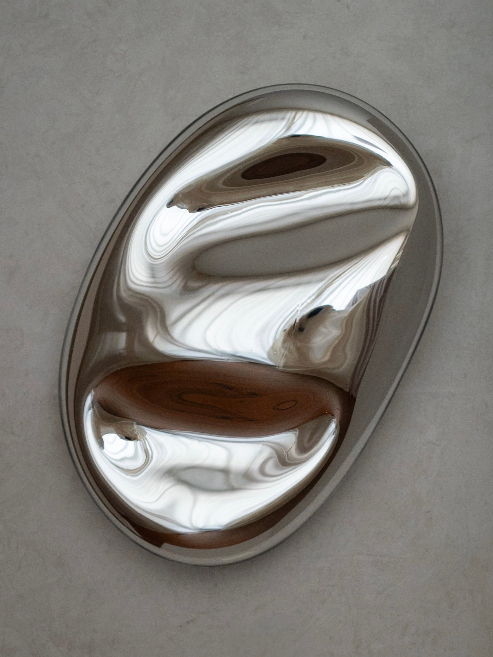The No. 46 Contemporary Mirror Sculpture Silver by STUDIO MILENA KLING is mounted on a light gray wall, featuring smooth metallic, oval-shaped reflective surfaces with wavy patterns that create an abstract design of light and dark tones, highlighting its handcrafted nature.