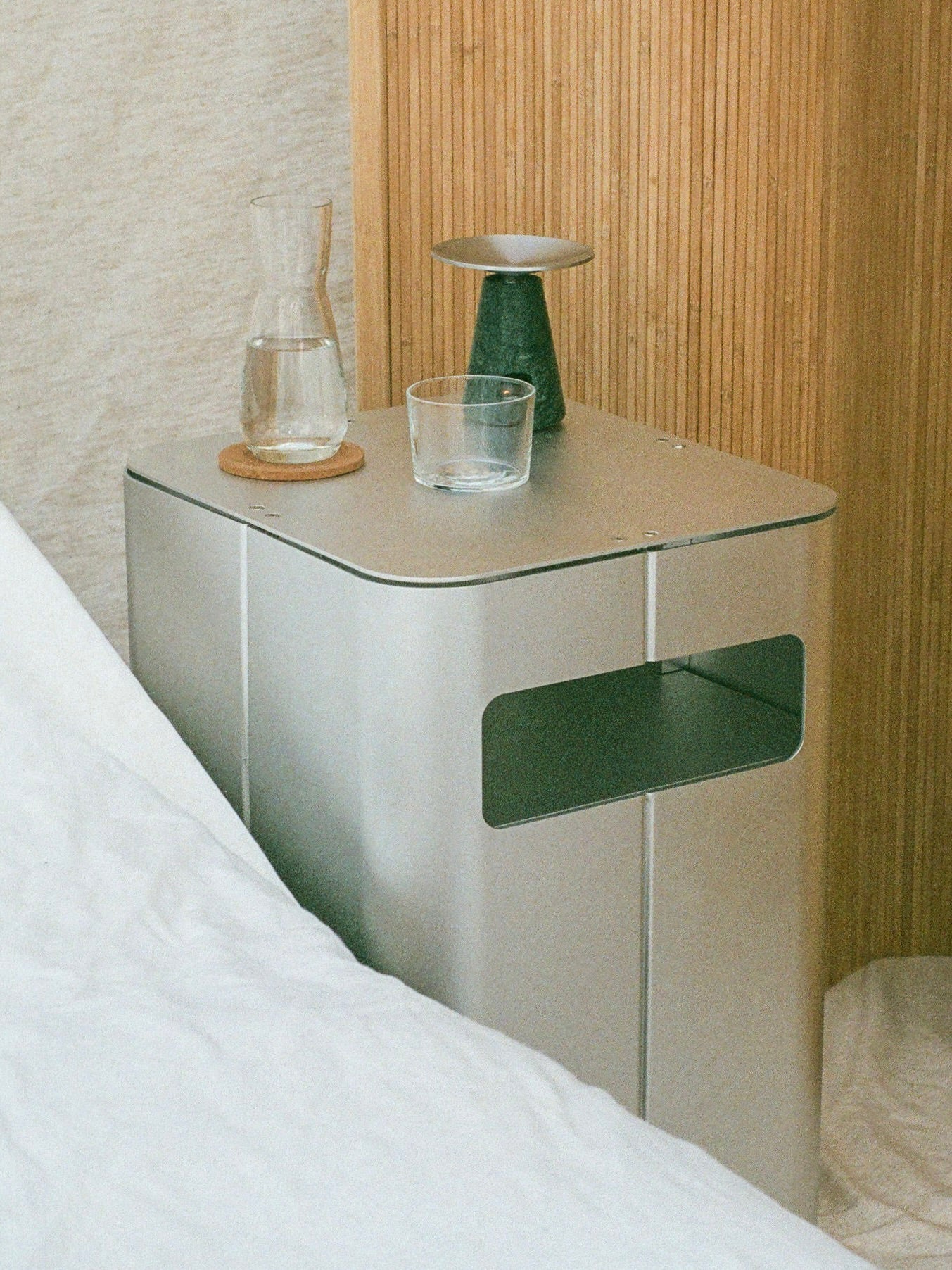 The 'Gadroon' Bedside Table by PARCTA, set against light wood paneling and a beige fabric wall, holds a glass of water, carafe on a coaster, small green vase, and glass dish for a sleek, functional bedside scene.