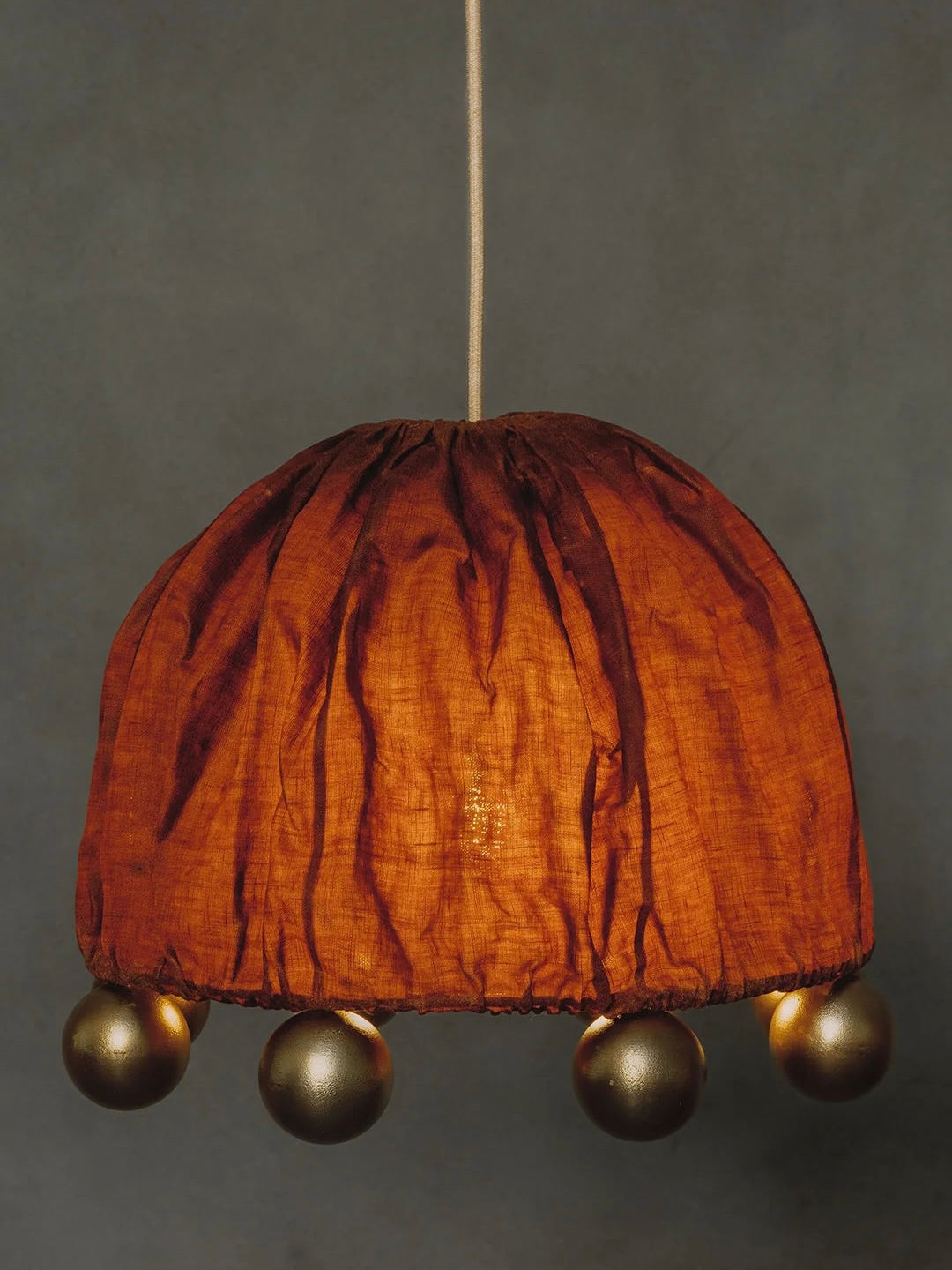 The "ALTER" pendant light by Kaytar showcases a round, orange beeswax linen shade with a wrinkled finish, accented by alchemy-inspired metallic spheres. A neutral gray background enhances its handcrafted charm.