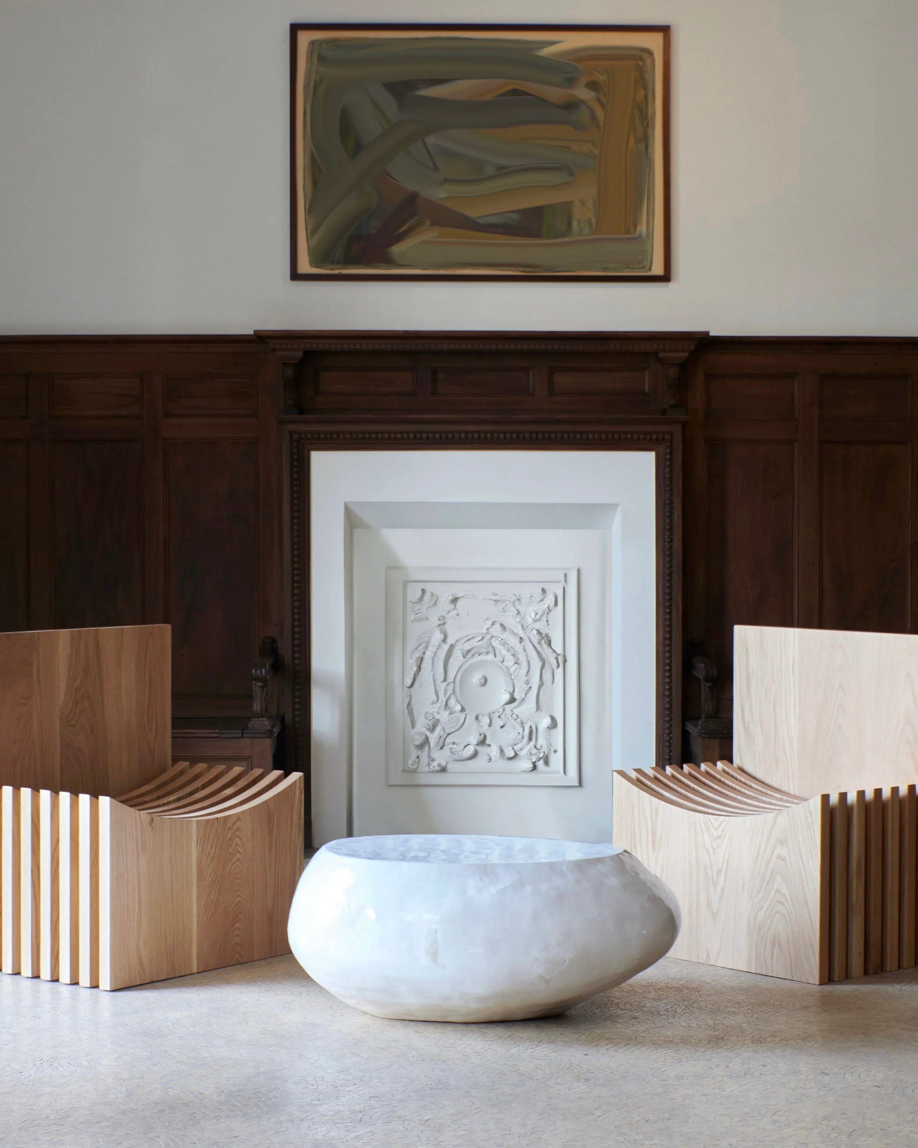 A minimalist room features two wooden chairs with slatted backs, a round Ceramic Coffee Table in Cream by Project 213A with customized glaze, and a large decorative fireplace with intricate molding. Above the fireplace hangs an abstract framed painting. The walls are partially lined with dark wood paneling.