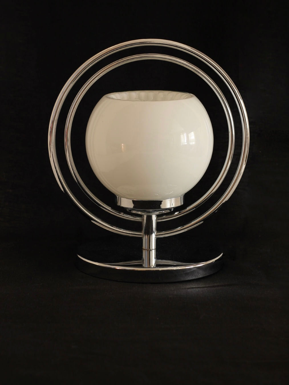 The Space Age Table Lamp from Boga Avante Shop boasts a white spherical lampshade encircled by two concentric metal rings. Its sleek chrome base enhances the mid-century modern design, beautifully contrasted against a black background.