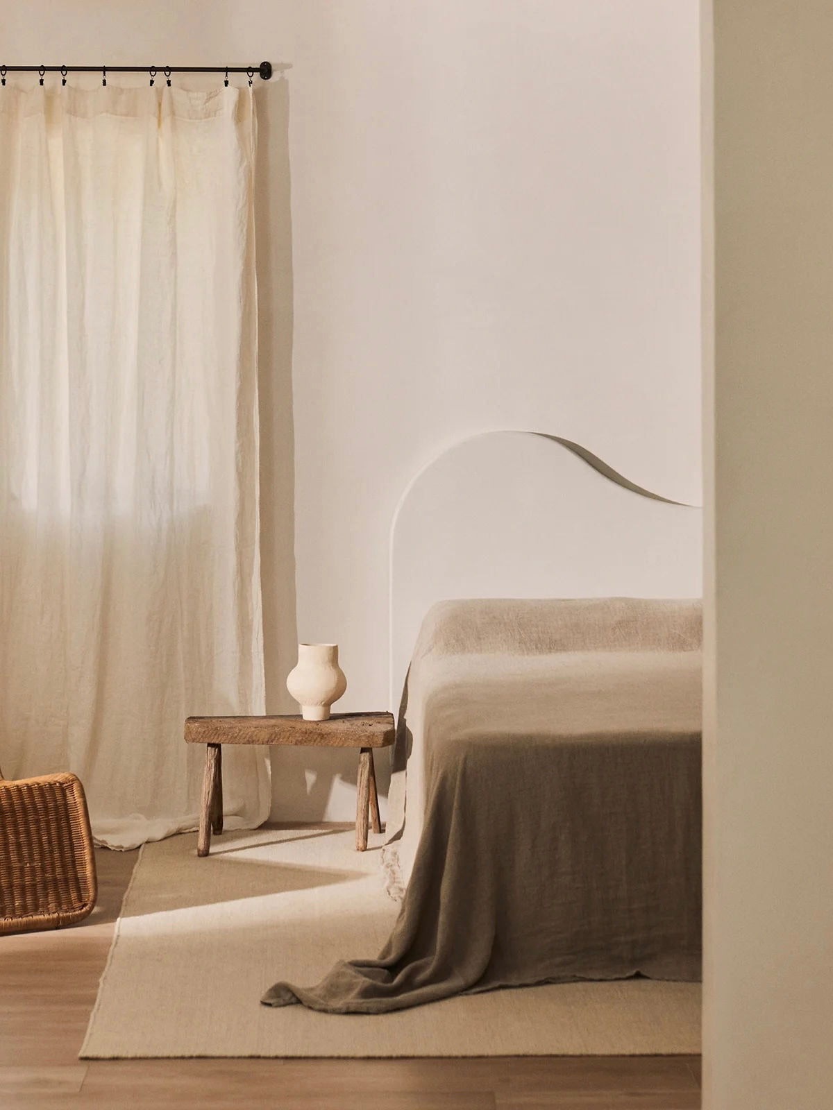 A minimalist bedroom features a Pasiego Headboard by MarlotBaus paired with a beige bedspread. A wooden stool holds a vase, while sheer curtains allow gentle light. A woven basket on the floor enhances the serene and cozy ambiance with handmade details.