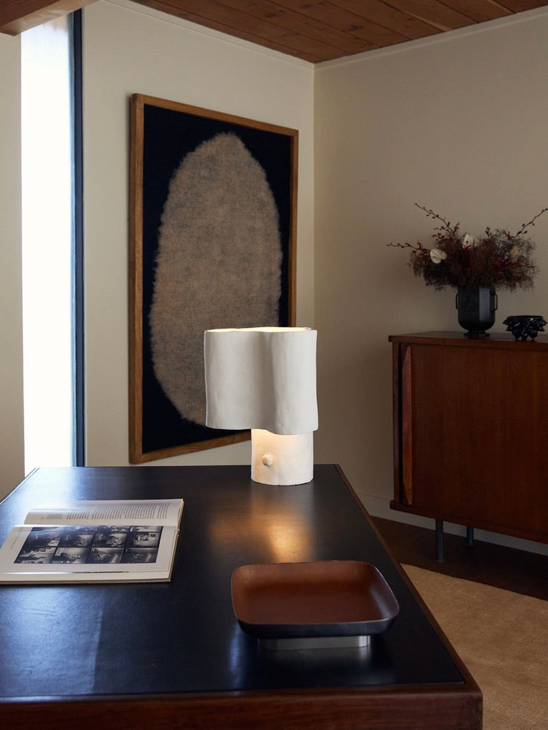 A modern interior showcases a black desk with an open book, a wooden tray, and the Cantilever Freeform Table Lamp by Studio Tristan Kallas. A large textured artwork graces the cream wall, while a floral arrangement adorns a handcrafted wooden cabinet, adding warmth to the space.