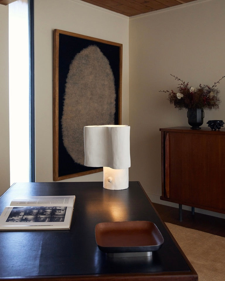 A modern interior showcases a black desk with an open book, a wooden tray, and the Cantilever Freeform Table Lamp by Studio Tristan Kallas. A large textured artwork graces the cream wall, while a floral arrangement adorns a handcrafted wooden cabinet, adding warmth to the space.