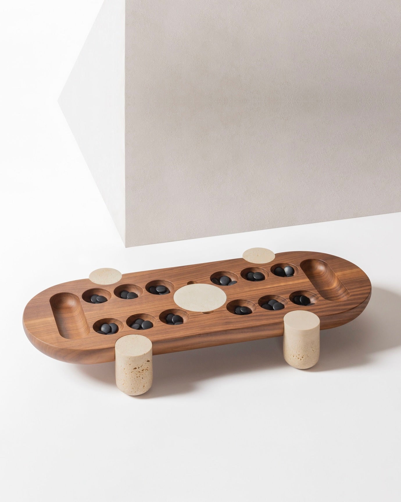 The MOCAMBO Wari Game Set in Wood and Stone by GIOBAGNARA features a solid walnut top with oval depressions for black stones, marble side legs, and is designed by Simone Fanciullacci, shown on a white background with angular walls.