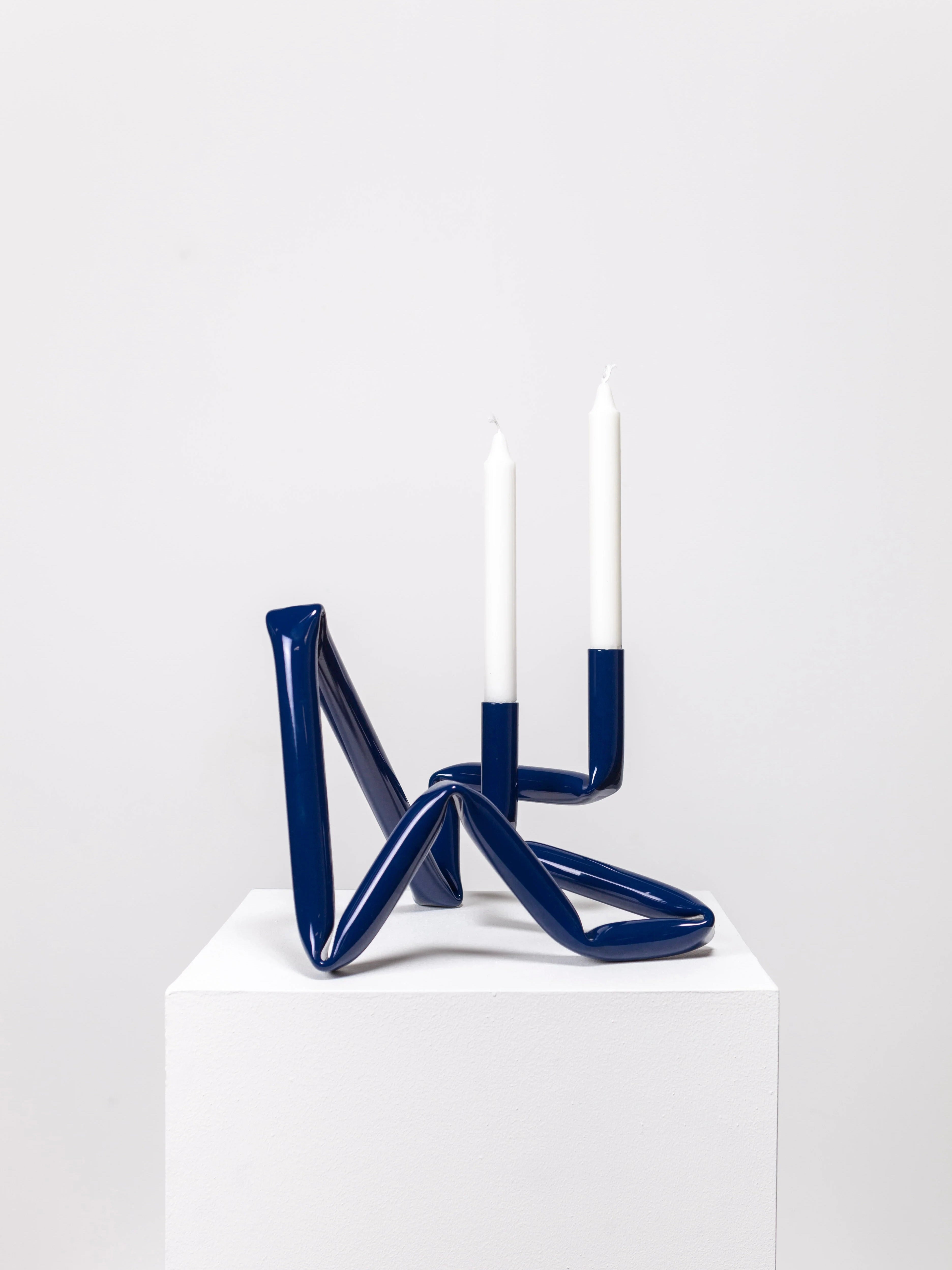 A modern, abstract Bucatini Candle Holder in Midnight Blue from AOT STUDIO, featuring two white candles. The handcrafted Denmark design boasts a unique, irregular looped structure crafted from sleek aluminum tubing and stands on a white pedestal against a plain white background.