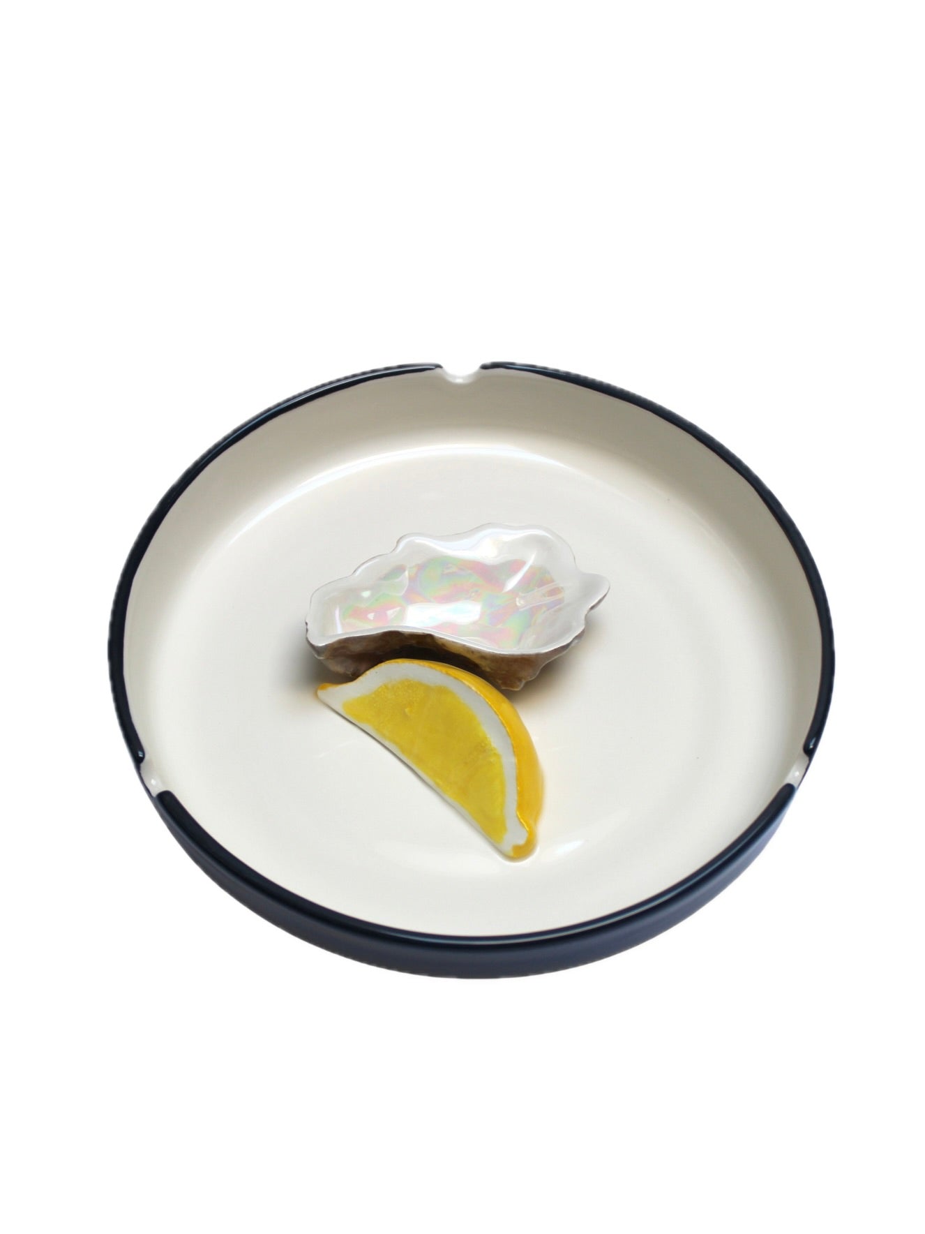 The Plaisir Solitaire Ashtray by Villa Arev is a refined white ceramic plate with a dark rim, featuring a small iridescent shell-shaped dish and a realistic-looking lemon wedge decoration.
