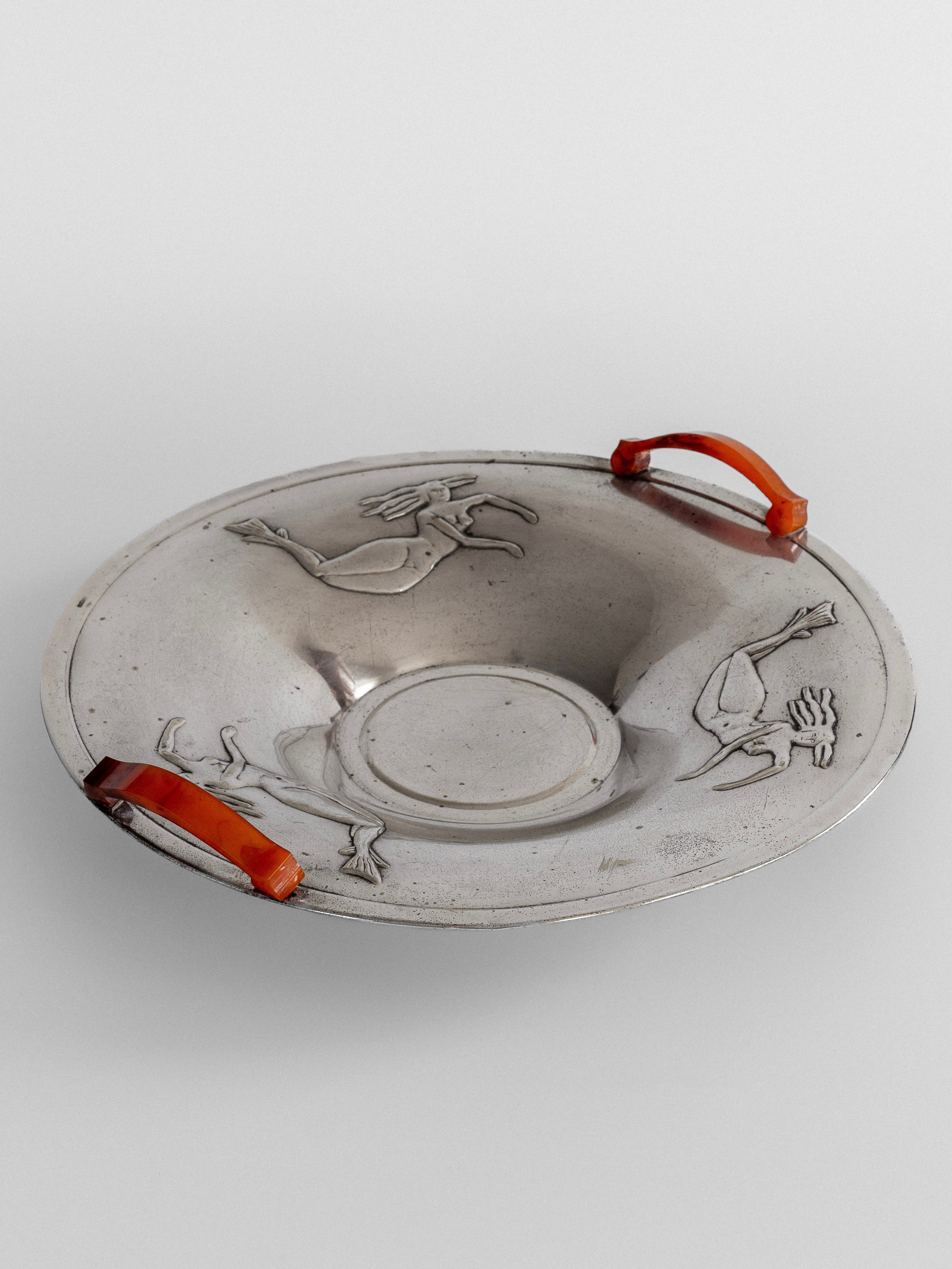 Introducing Spigel's Decorative Bowl with Mermaids from the 1930s: a silver-plated masterpiece adorned with two vibrant orange handles on opposite sides. This bowl, reminiscent of the Swedish Grace design, showcases engraved images of two figures in motion and features a central circular indentation.