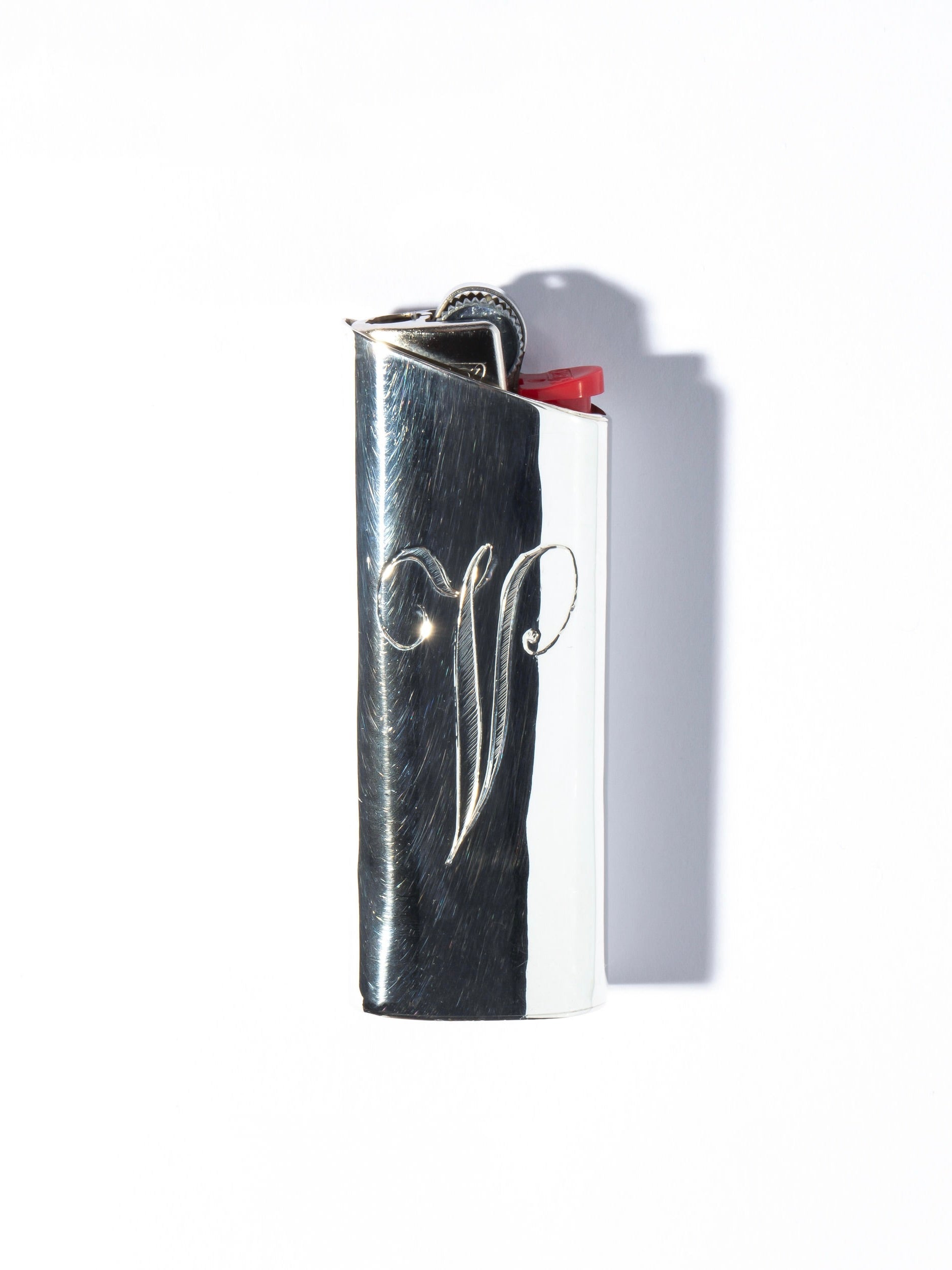 The Pedro Ventura Monogram Lighter Case Maxi Size is a metal lighter with a hand-engraved, shiny sterling silver case showcasing a decorative design on the front and a red ignition lever at the top. Shadows beneath indicate it's resting on a flat surface.