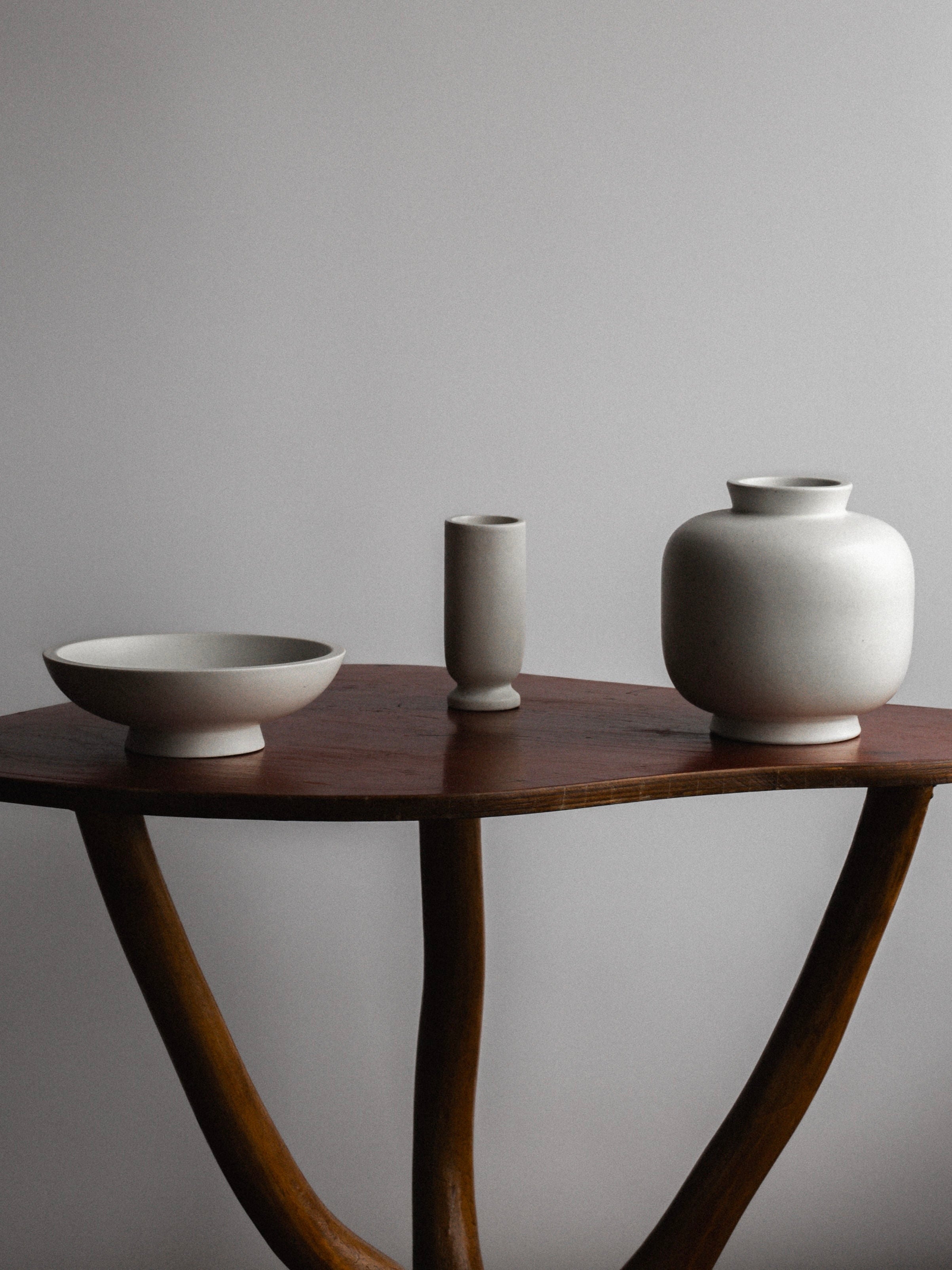 A minimalist setup on a rustic wooden table features three white ceramic items from Spigel, named the 'Carrara' Ceramic Trio, 1940s, Sweden: a shallow bowl on the left, a slender cylindrical vase in the center, and a round jar with a narrow opening on the right. The subtle Carrara glaze enhances their elegance, highlighting the simplicity of the scene.