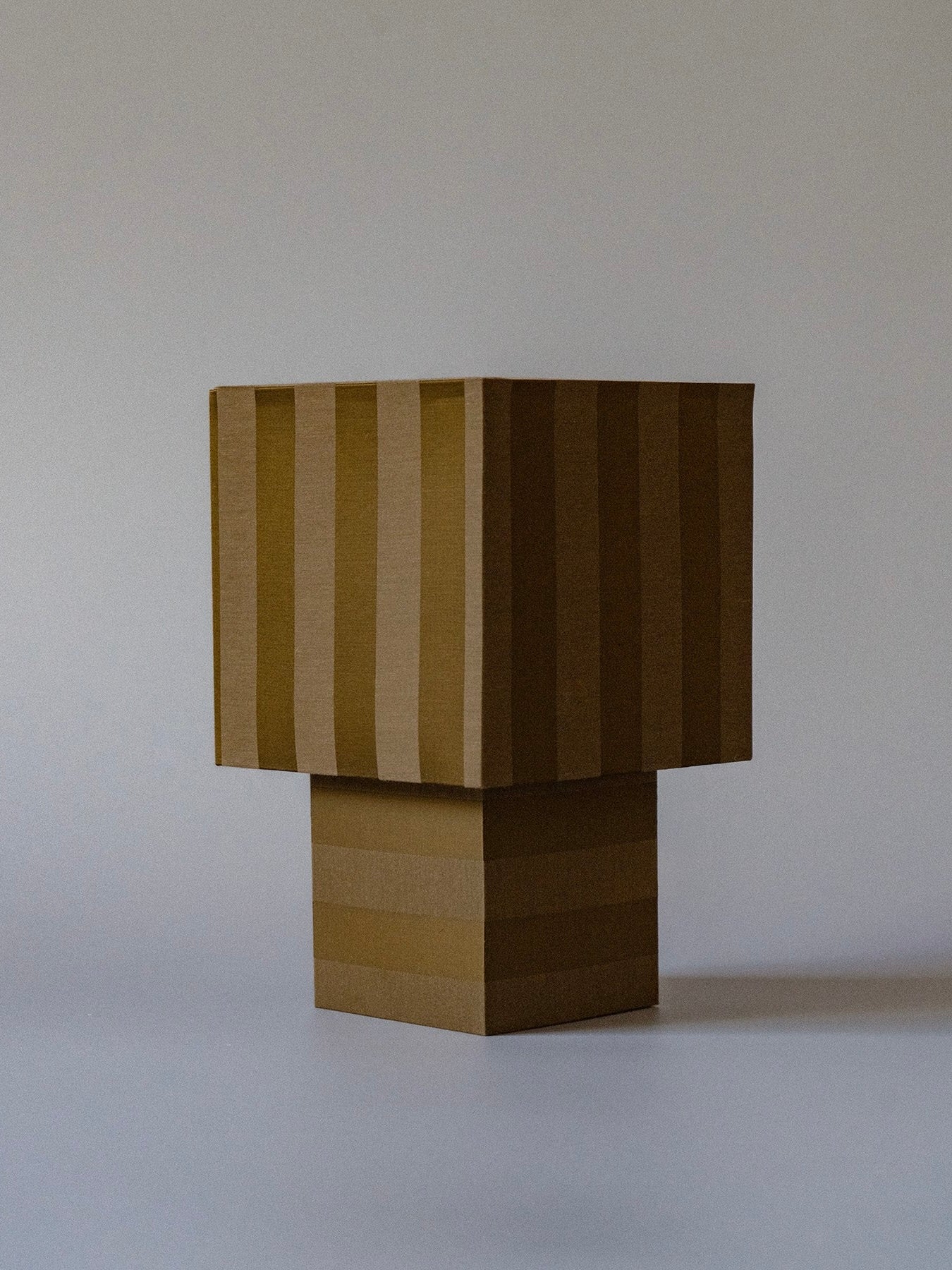 La Petite BoƮte" - Julii Table Lamp by mattina moderna is presented in a rectangular, vertically-striped cardboard box with alternating light and dark brown stripes against a light blue background. The box, exuding a touch of handcrafted playfulness, appears to be partially opened.