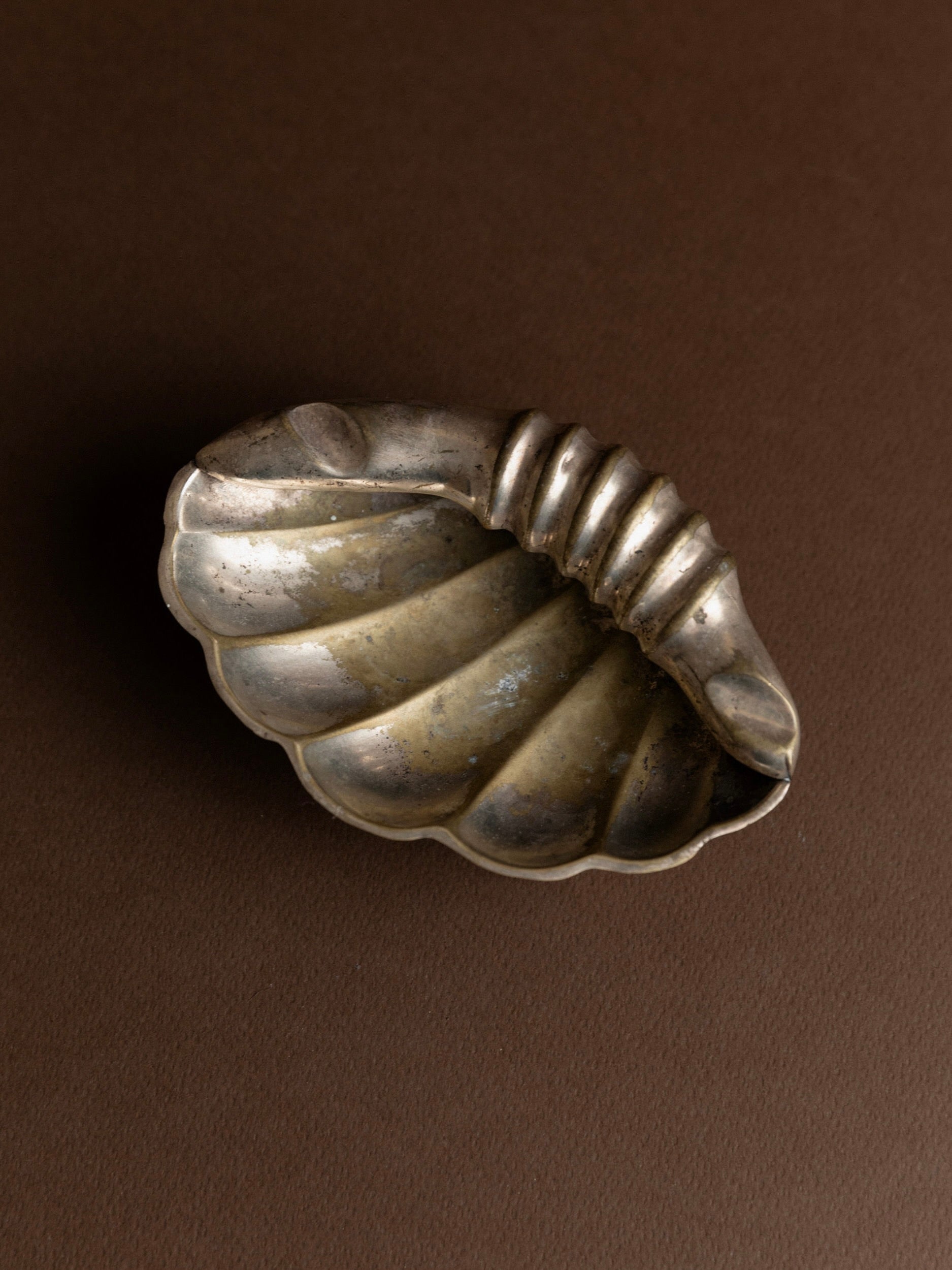 An Ashtray in Shell Shape from Bottega Jacobs, dating back to the 1960s in Italy, graces a brown surface. It features a ribbed, curved handle and boasts a tarnished patina that accentuates its intricate details and texture.