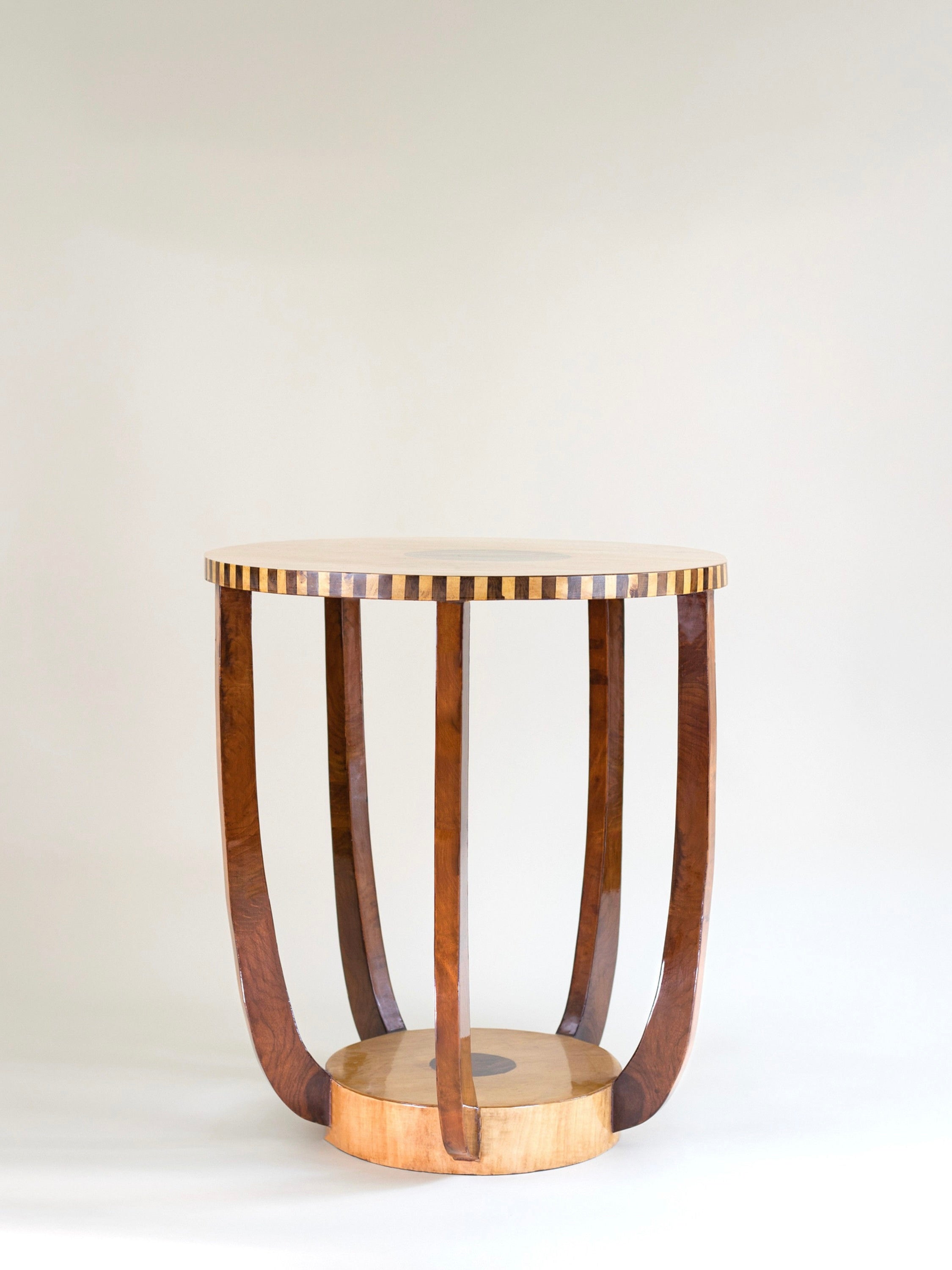 The 'Roman' Side Table by Meola Interiors is a mid-century Art Deco piece with a round top featuring an alternating light and dark wood pattern. Its burl wood veneer and elegant curved legs connect to a circular base, showcasing exceptional craftsmanship against a plain background.