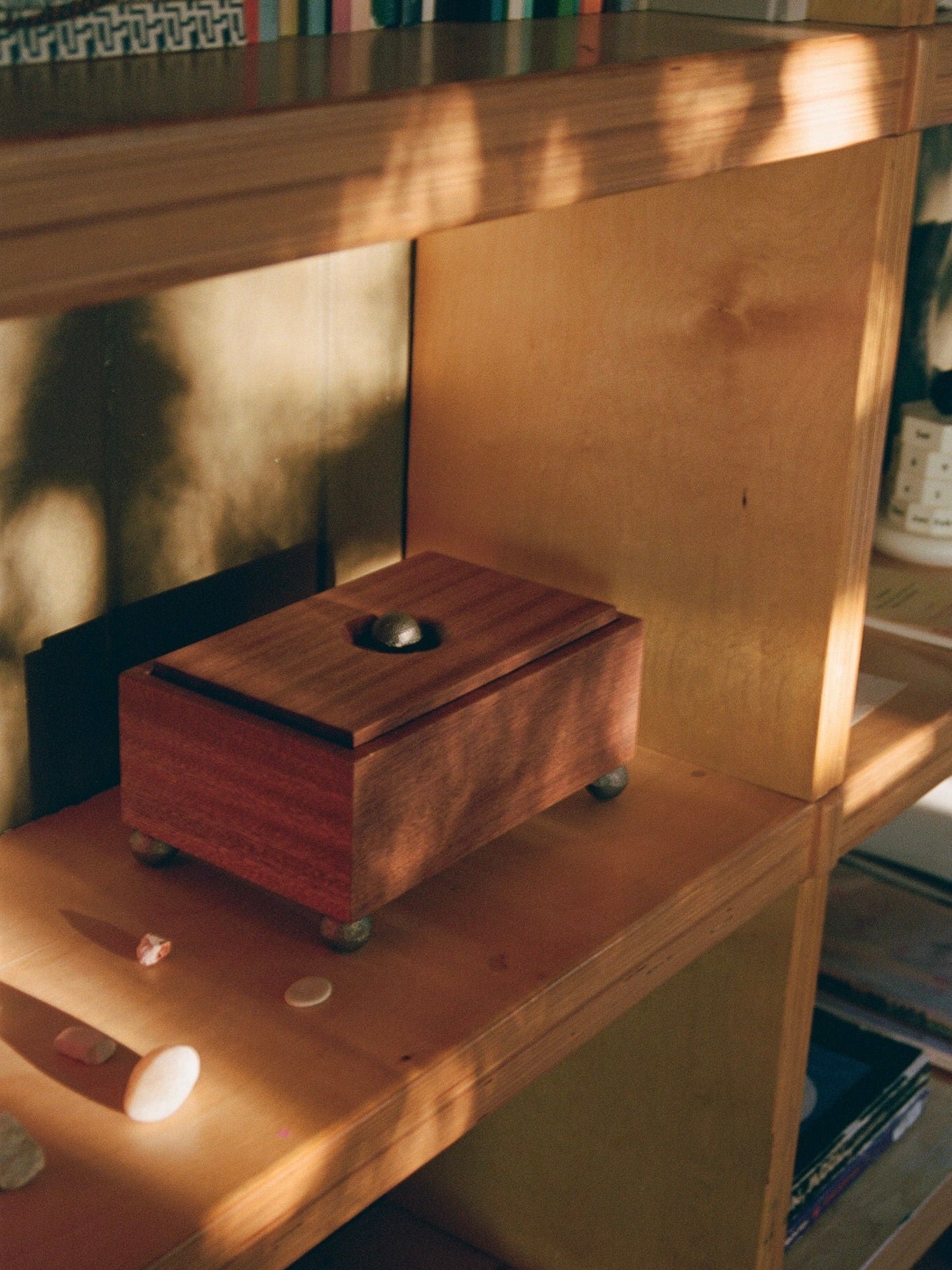 A wooden shelf bathed in dappled sunlight holds The Phone Box by MUHLY, a vintage wooden box with a round knob on top, suggesting a sense of phone-free living. Surrounding the box are small stones and objects. The shelf has other items, including books, but they are partially out of the frame.