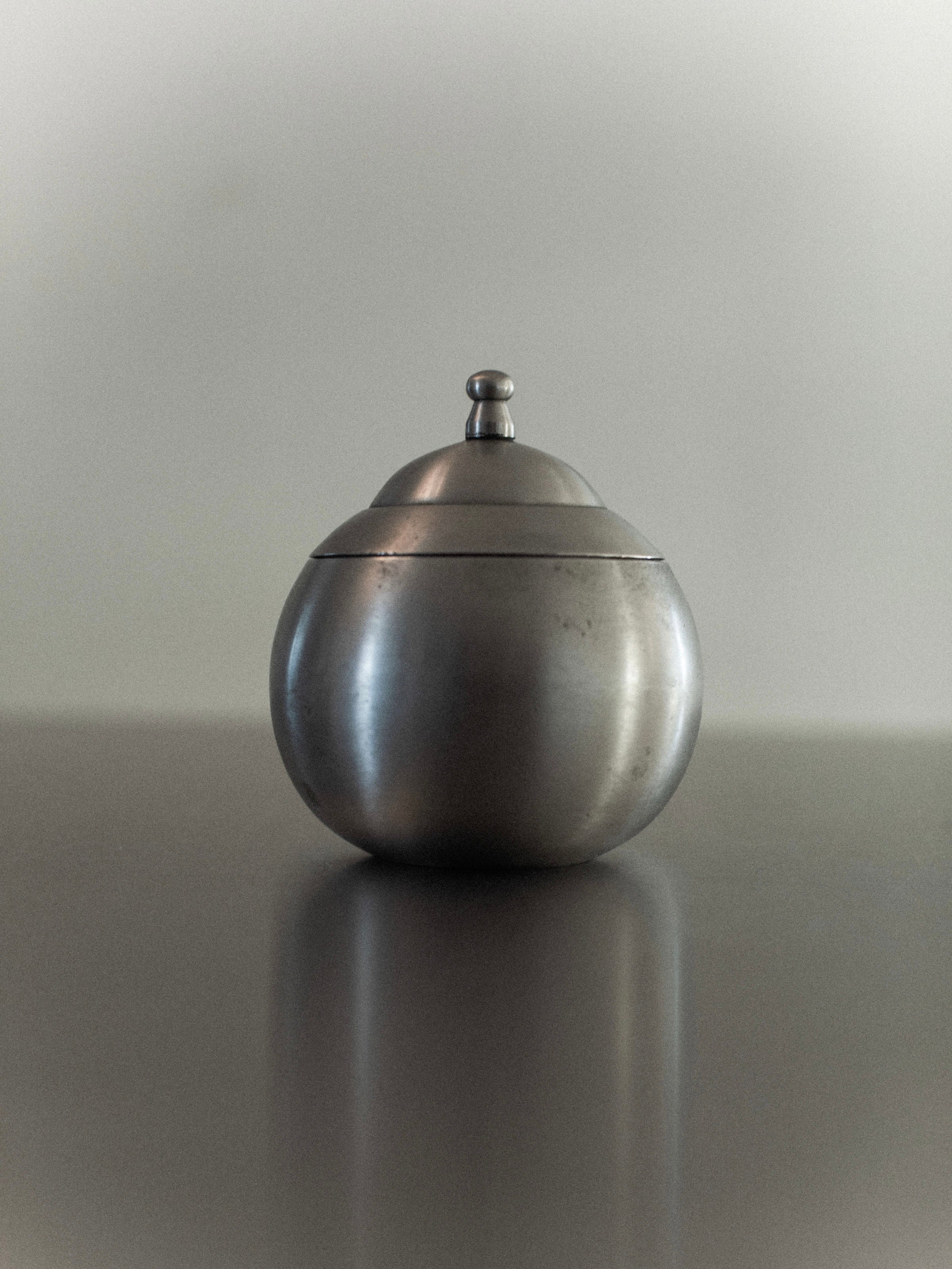 The Steel Bonbonnière by Out For Lunch is a vintage-inspired, round container crafted from silver-colored metal with a polished steel finish. It features a lid topped with a small knob and is elegantly displayed on a reflective surface against a neutral background, exuding industrial charm.