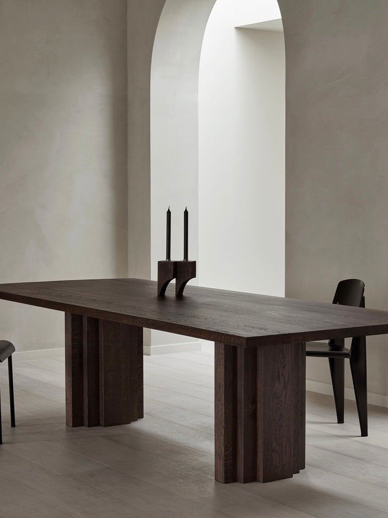 A minimalist room showcases a Mokko Brut Slim table made from European oak, accented with two black candles in a holder, and flanked by sleek black chairs. A white arched doorway and light-colored floors and walls enhance the space.