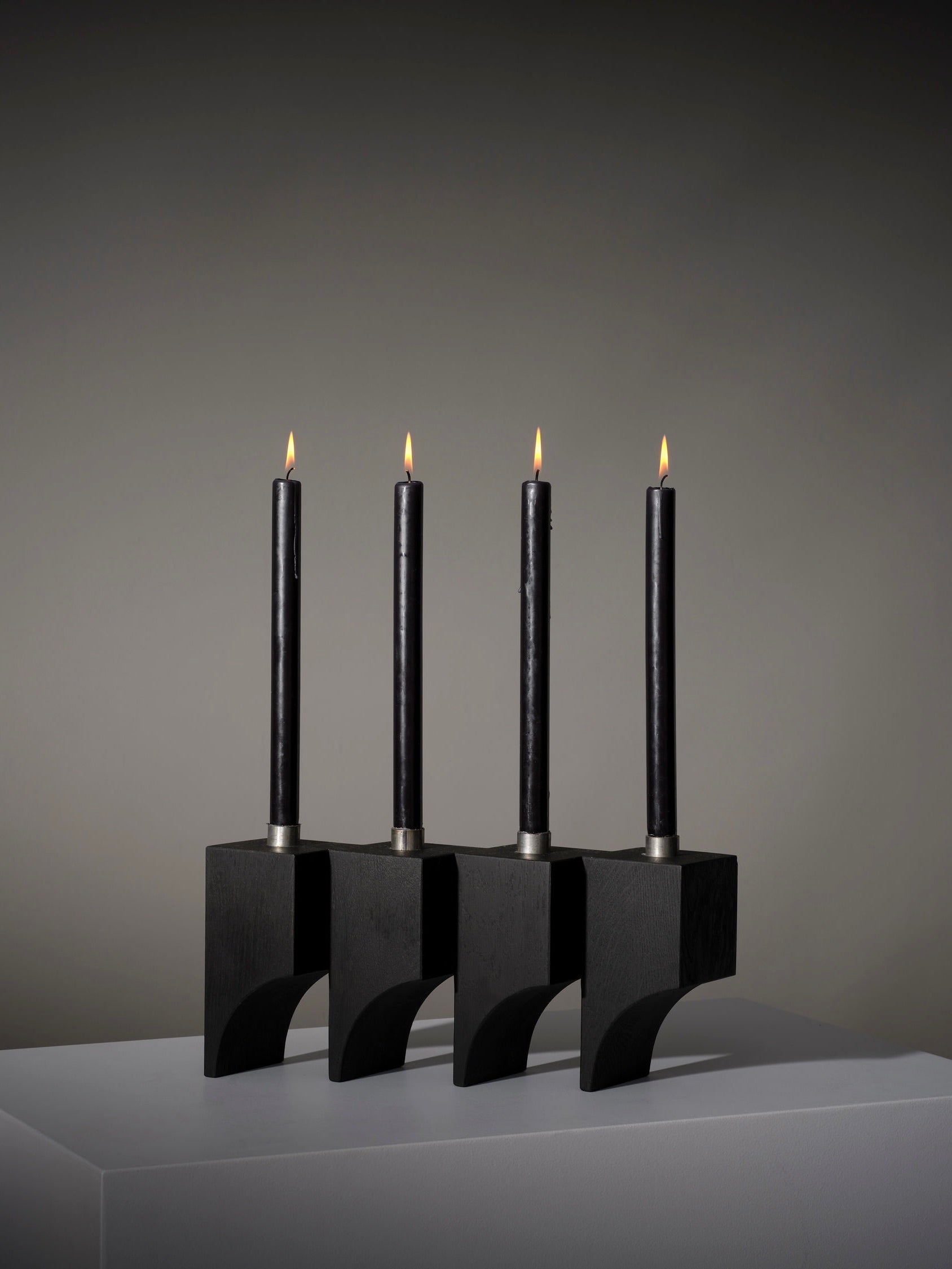 Four lit black candles sit in a sleek Acer Candle Holder R:4 by Mokko, featuring a geometric design against a neutral gray background. The setup mimics architectural cubic volumes and rests on a white surface, creating a minimalist look.
