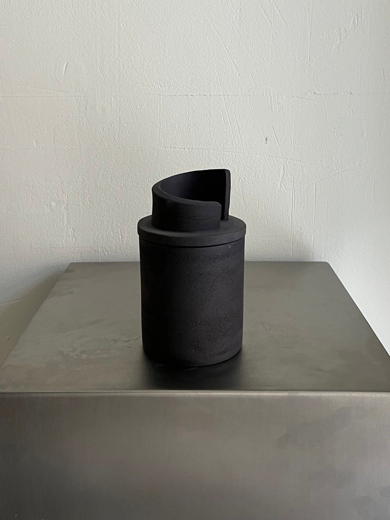 The Sculptural Scented Candle by 2222Studio, with a unique rounded top, rests on a metallic surface against a plain white wall. This limited edition matte black piece showcases modern minimalist design, contrasting sharply with the smooth reflective table below.