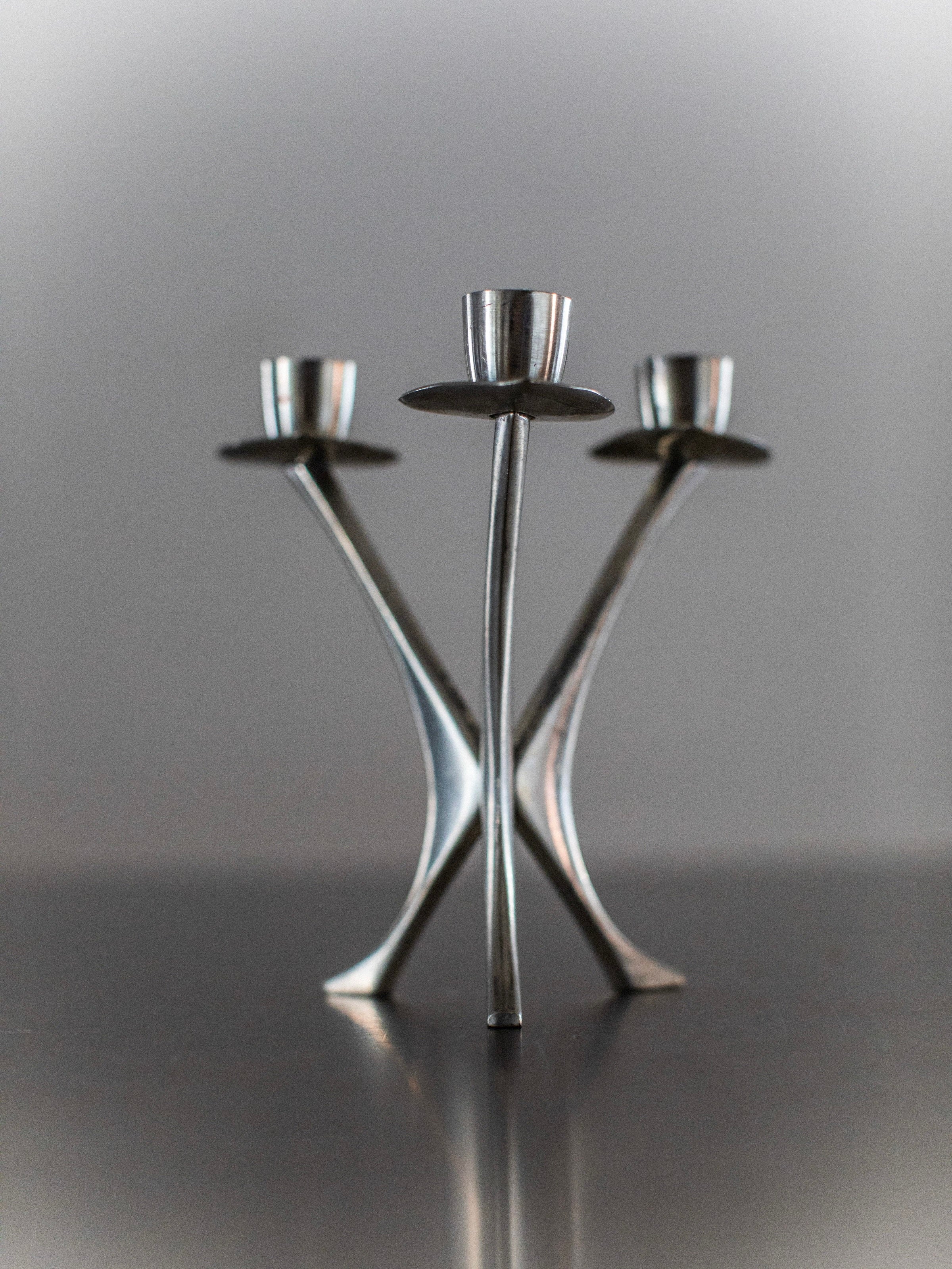 The Out For Lunch Steel Candleholder is a sleek, modern decor piece featuring three upward-facing holders. With its curved, intersecting stems set against a neutral gray background, it exudes an artistic and minimalist elegance.