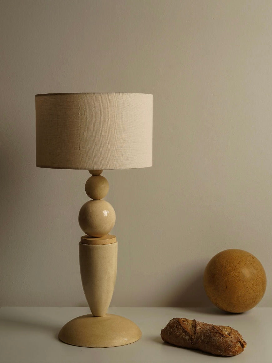 A beige Fauvery 70's Ceramic Table Lamp from the Les Objets Amsterdam collection, showcasing a modern geometric design with stacked spherical and oval shapes as its base, sits on a white surface. Beside the lamp are a rustic loaf of bread and a yellow spherical object, all set against a neutral-colored wall.