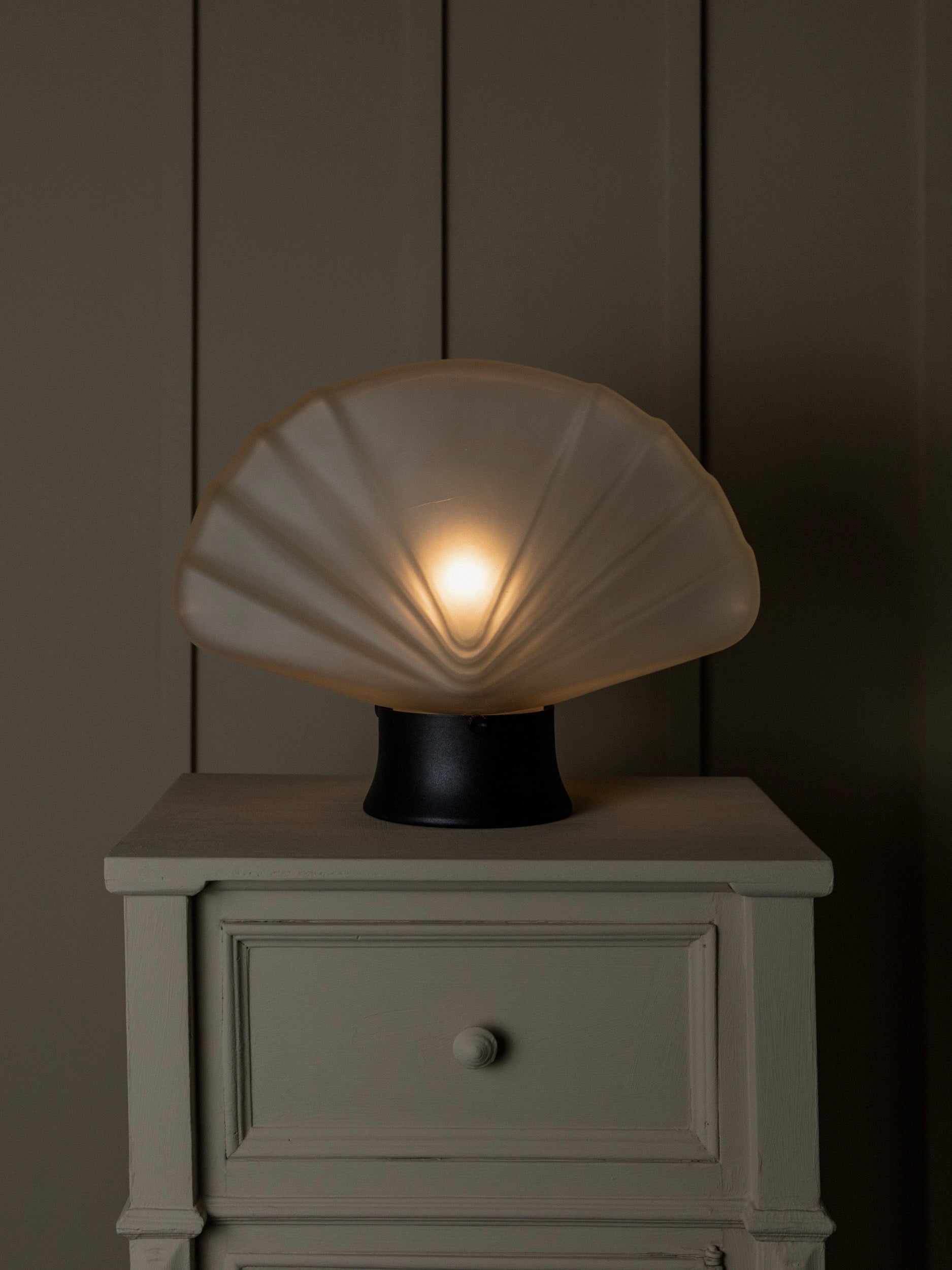 A Murano Glass Table Lamp 80s by Bottega Jacobs casts a soft glow on a wooden nightstand against a dark beige wall. Its translucent shell design creates a cozy ambiance, blending elegance and warmth perfectly.
