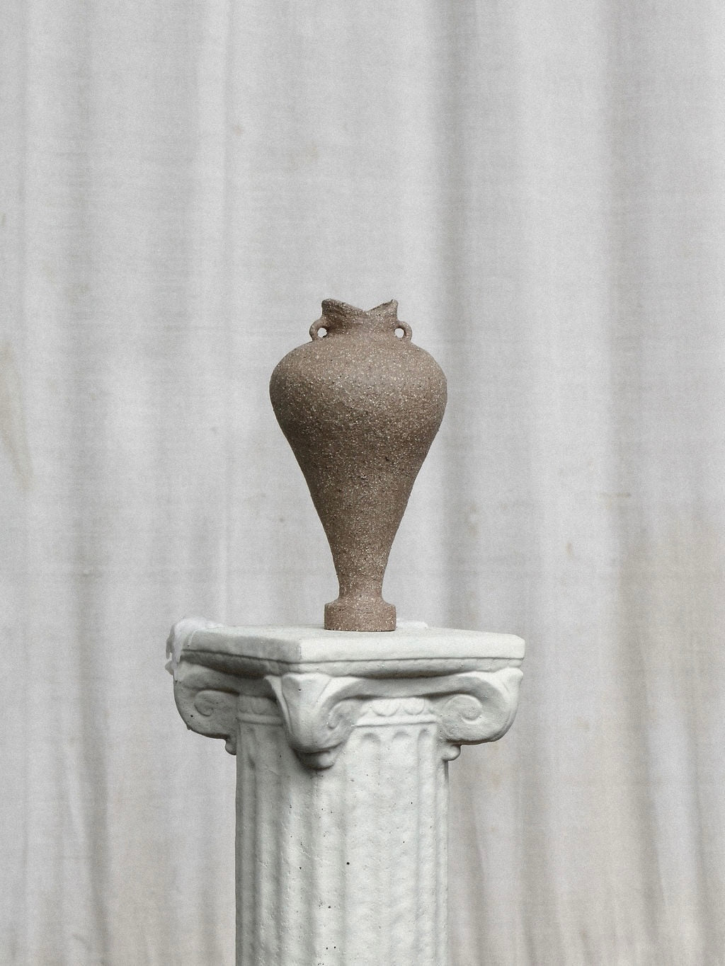 An Amphora Vessel by Emma Lindegaard Studio, featuring a textured, ancient-style design and small handles crafted using the coiling method, rests on a decorative white column against a plain gray backdrop.