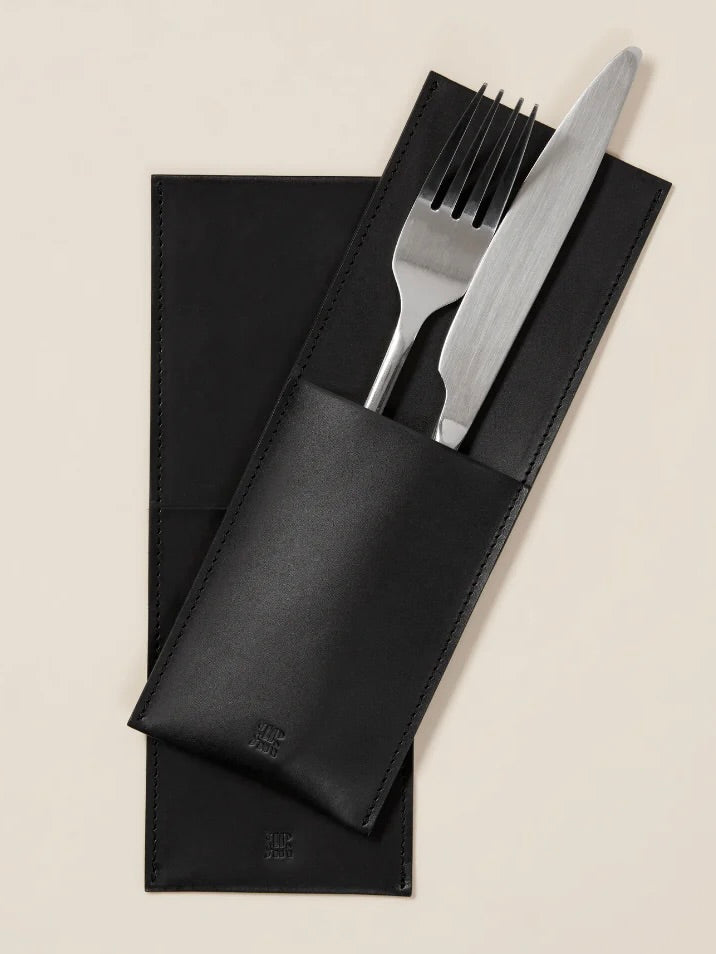 A stainless steel fork and knife sit inside a Leather Cutlery Holder from Paradise Row, crafted from vegetable-tanned leather, partially open to showcase the cutlery. The holder is on a matching black leather placemat against a beige backdrop.