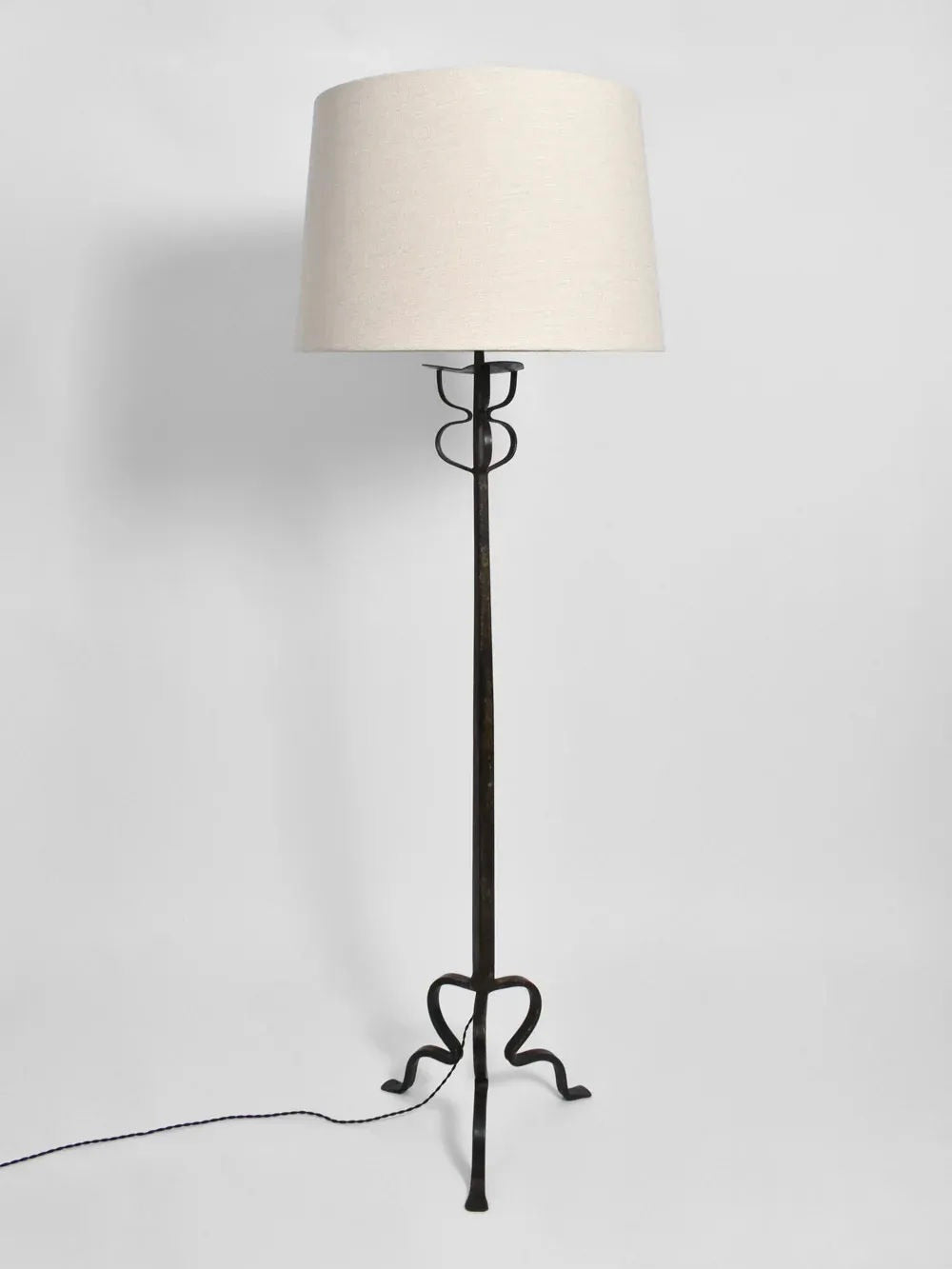 The Tripod Iron Floor Lamp from Barracuda Interiors features a tall, slender stand with a graceful curved design at the base, crafted in wrought iron. It is topped with a beige cylindrical lampshade and has an elegantly extending black power cord, reminiscent of 1960s France.