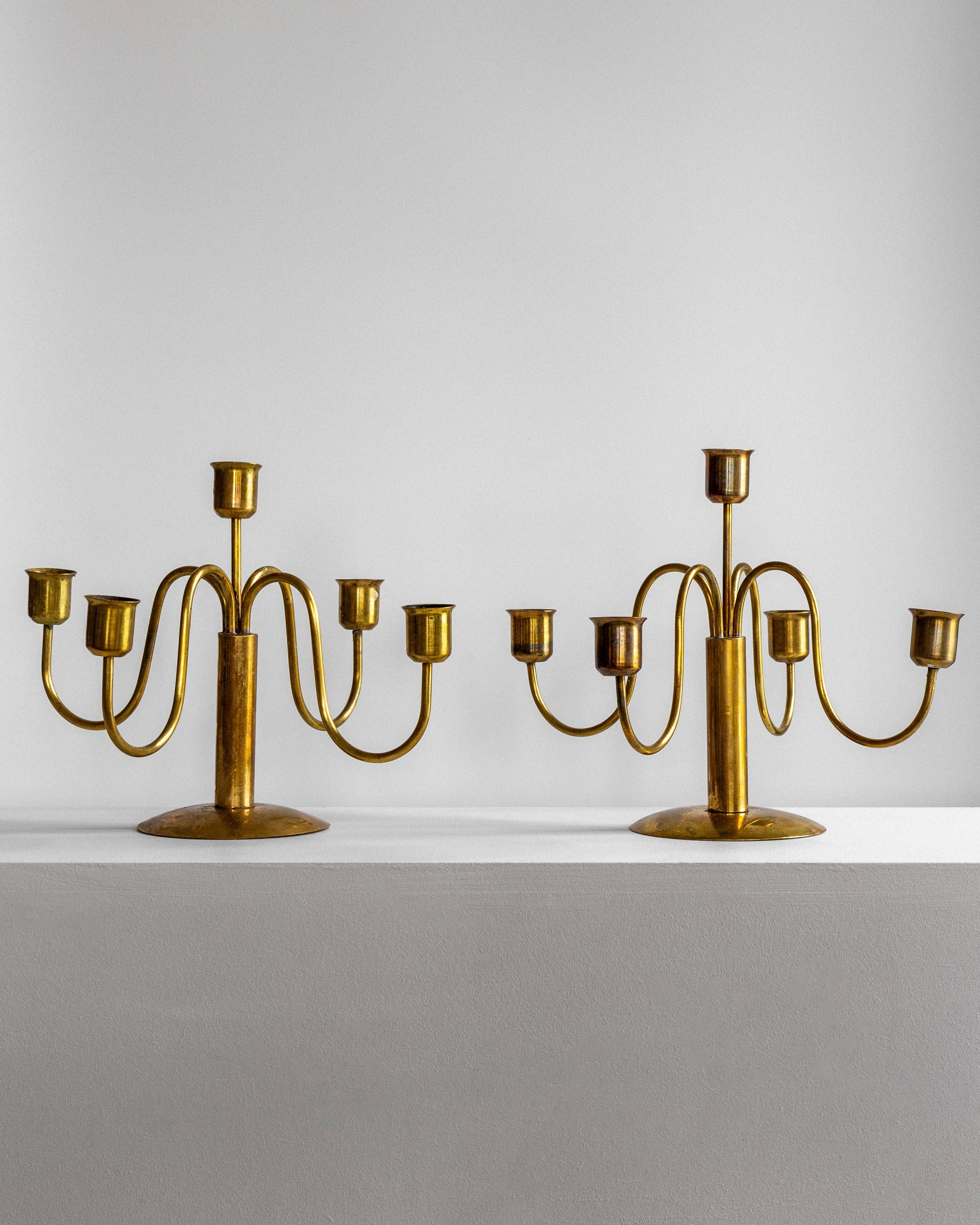 Two Spigel Mid-Century Brass Candelabras, each featuring six arms and candle holders, are showcased on a white surface against a gray background. The symmetrical and elegant design boasts ornamental details with a classic, antique appearance. A subtle patina enhances their timeless charm.