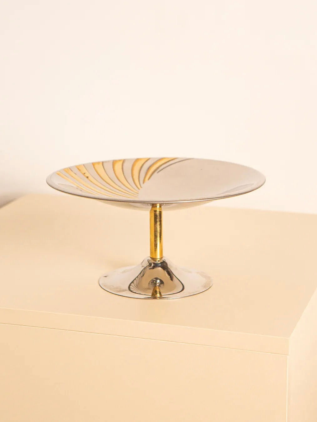 A charming retro piece, the Treaptyque 60's Fruit Coupe in Metal and Brass features a clear glass pedestal dish with a gold swirl design on the plate, elegantly displayed on a light beige surface against a plain white background.