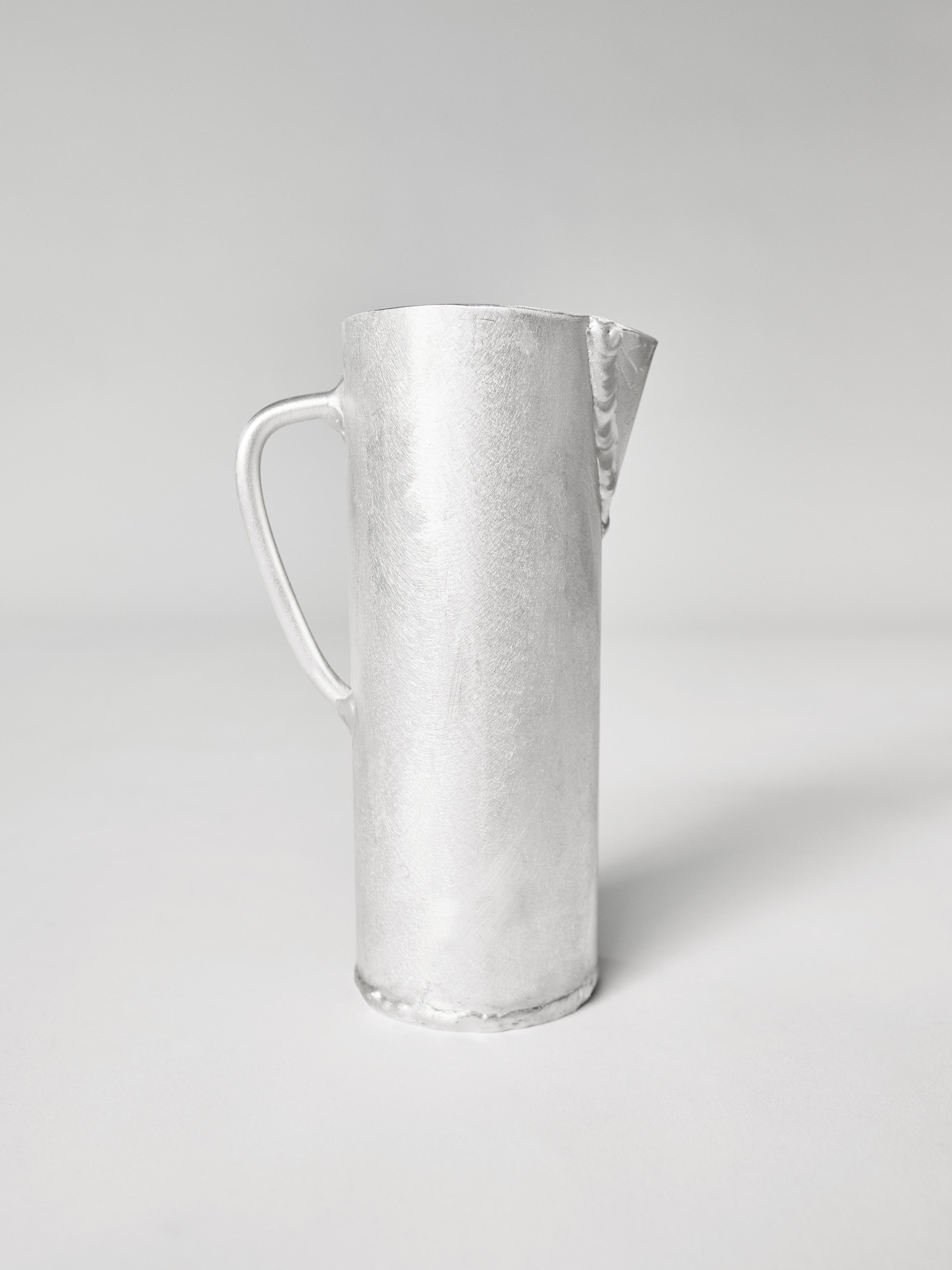The Aluminum Jug by Six Dots Design is a silver pitcher with a minimalist design, showcasing a cylindrical body, small spout, and curved handle. Handcrafted in a North London studio, its simplicity is accentuated against a plain light gray background.