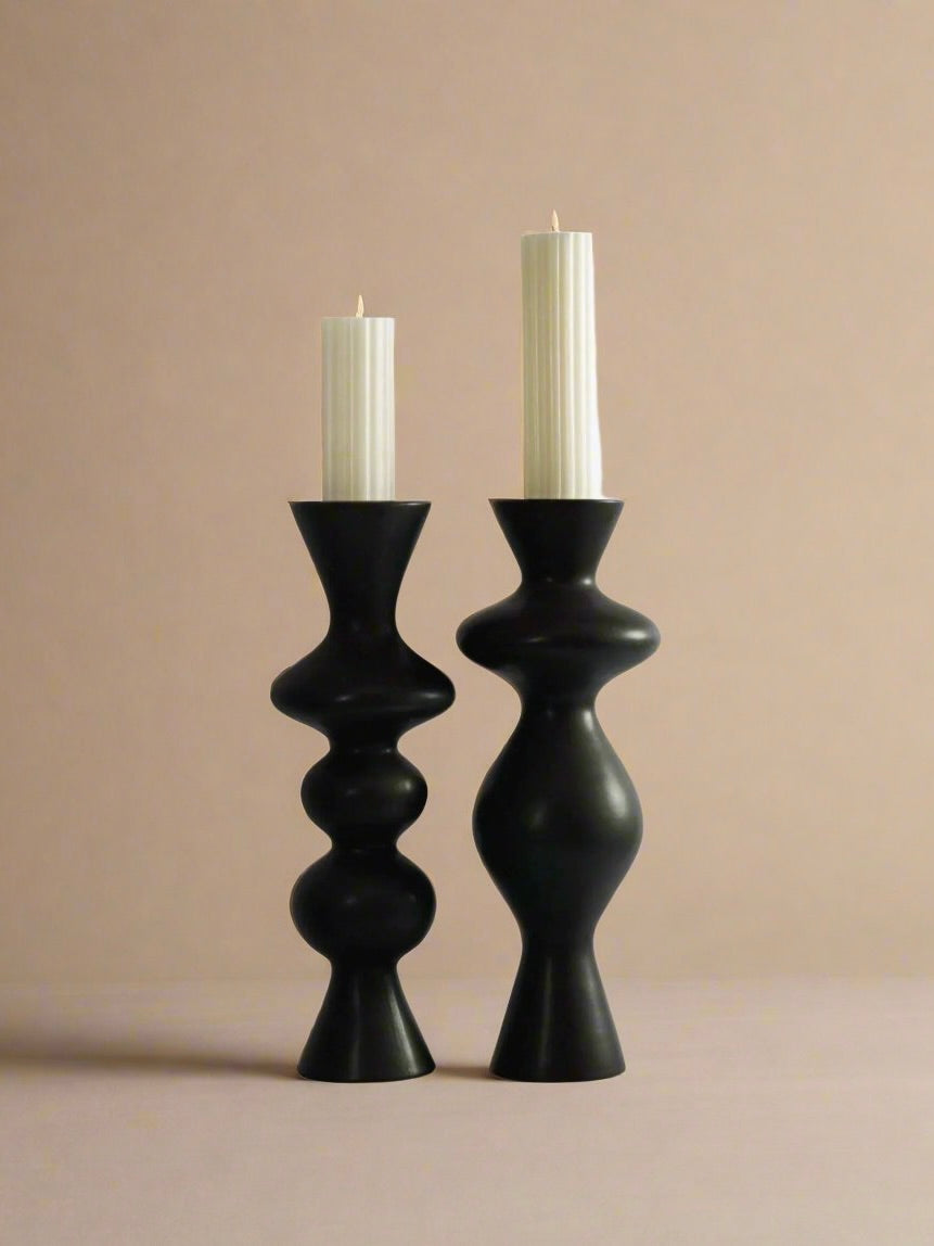 Two lit white candles are showcased in Ransom & Dunn's Pair of Ceramic Candleholders, featuring modern black designs that mimic curvy, sculpture-like shapes reminiscent of the female form. Set against a soft beige background, these candleholders vary slightly in shape, adding intriguing visual interest.