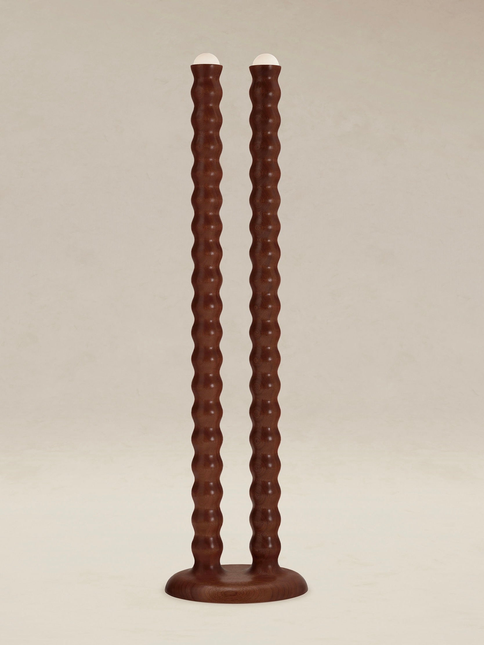 Introducing the "Tougarine" Floor Lamp by AÈDE Studios, featuring a pair of tall, wavy brown shafts crafted from cognac-red mahogany wood. These lamps elegantly stand upright on simple round bases with white tops, set against a neutral background.