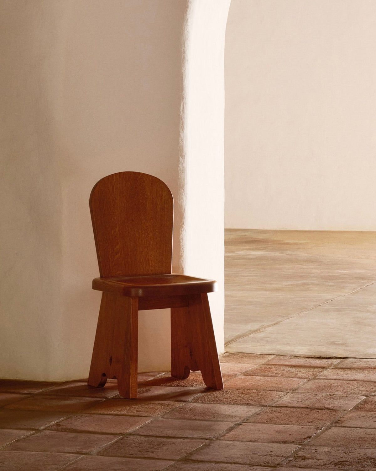 A Honey French Oak Wood Rambling Chair by Lemon sits against a white, textured archway wall on reddish-brown tiles. The warm minimalistic atmosphere with soft natural lighting suits contemporary living perfectly.
